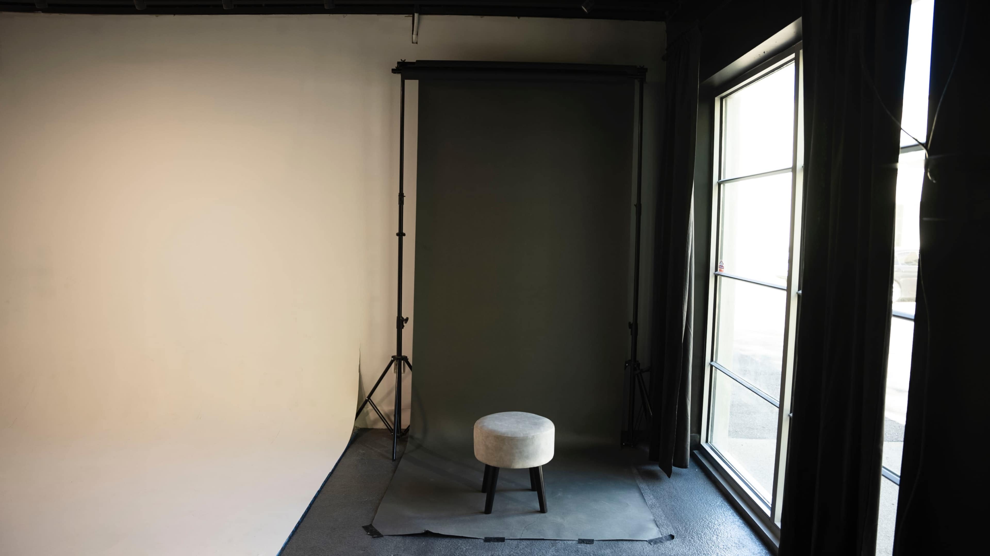 An empty photography studio with a gray backdrop, a stool in front of it, and a large window letting in natural light.