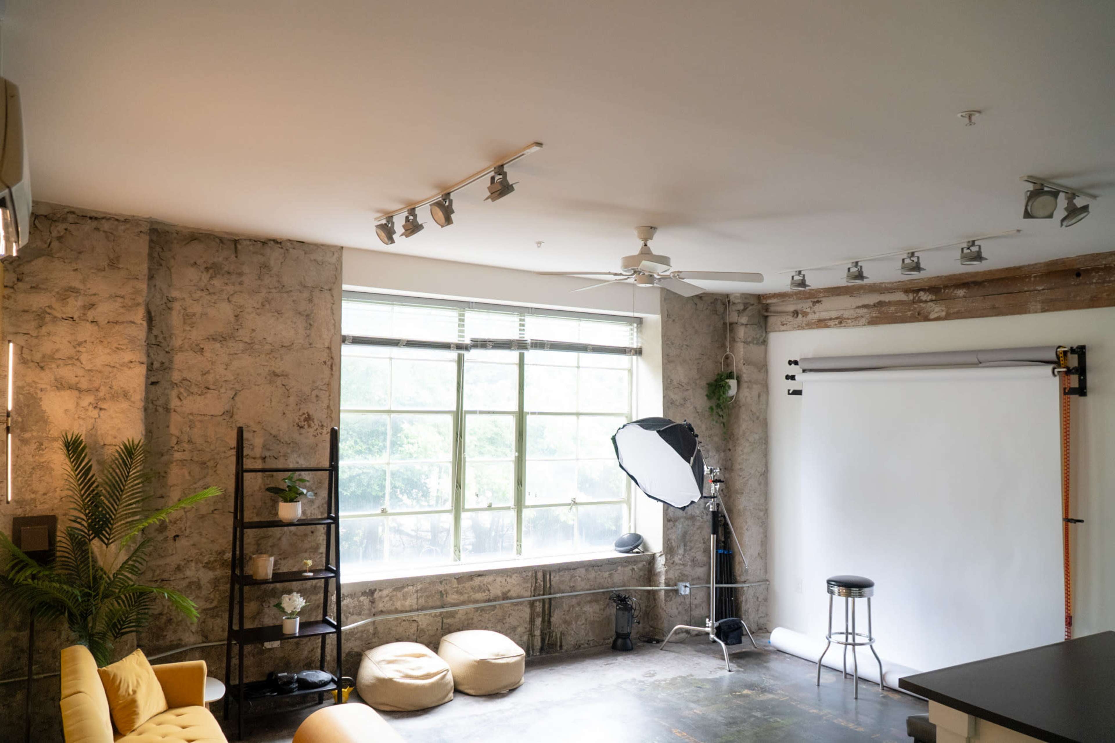 The image shows a photography studio featuring a large window, beige bean bag seating, a black stool, and a backdrop setup with a softbox for lighting.