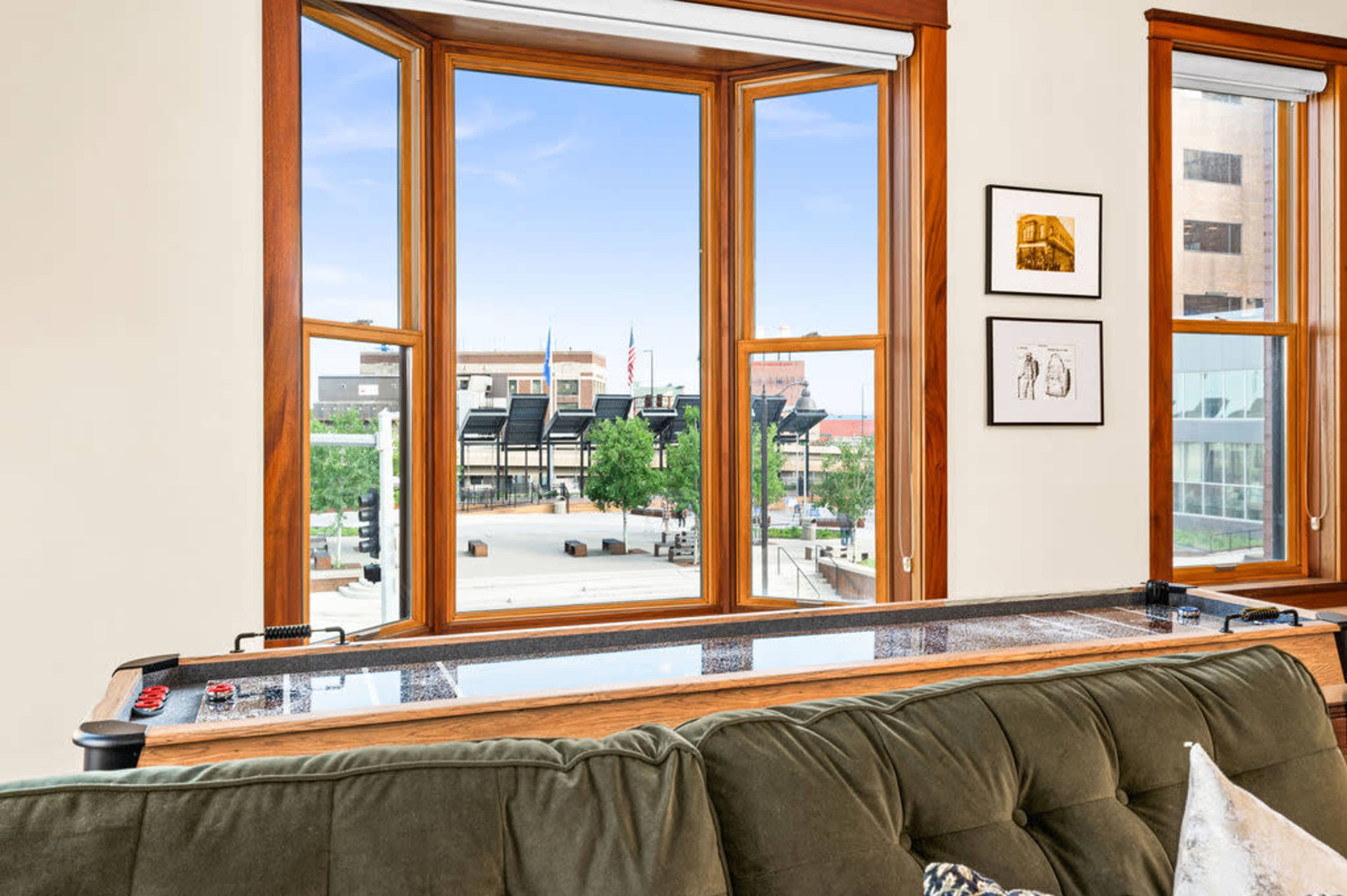 A view through large windows shows a recreational area with greenery and buildings in the background, featuring a game table in the foreground.