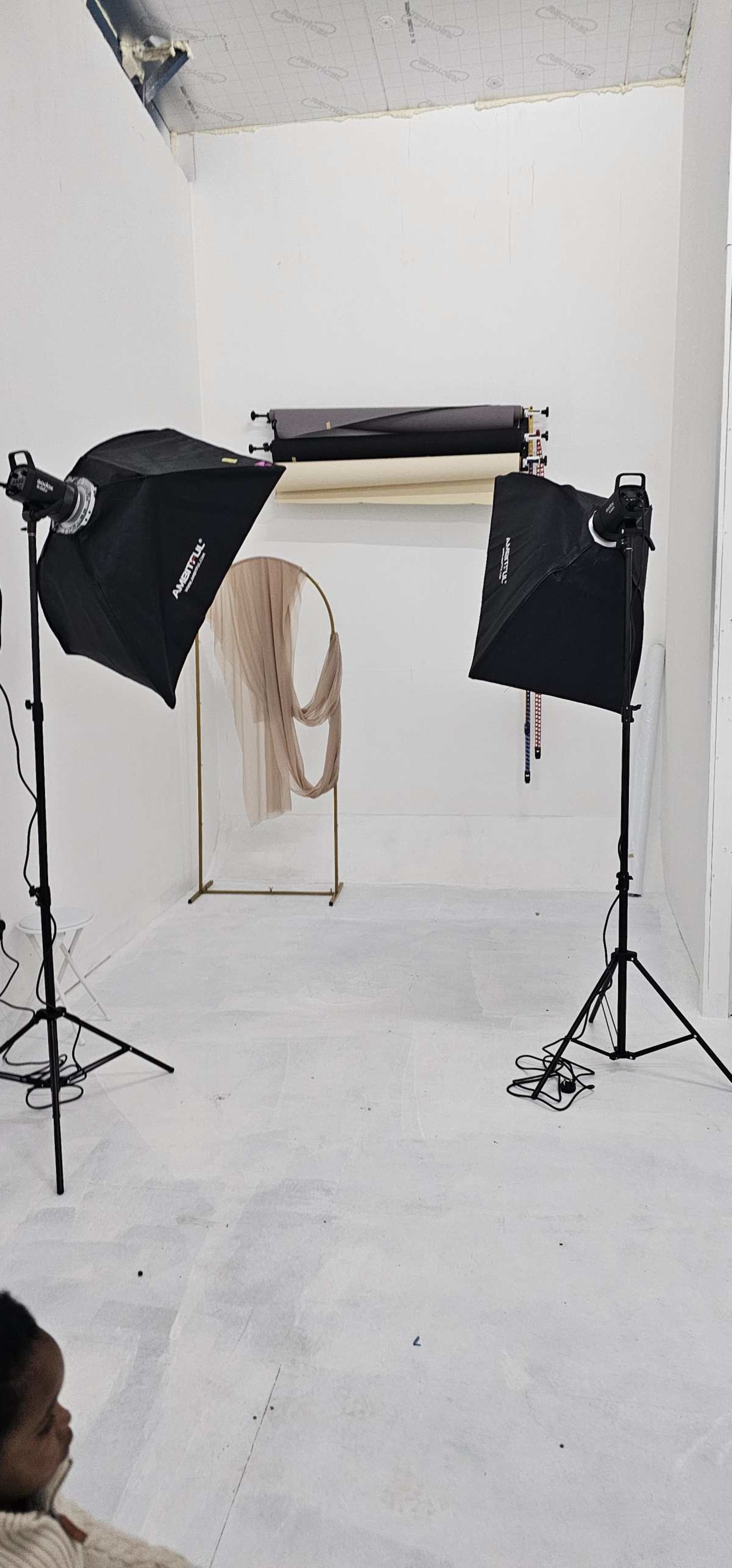 The image shows a photography studio with two softbox lights, a backdrop stand with drapes, and a white floor.