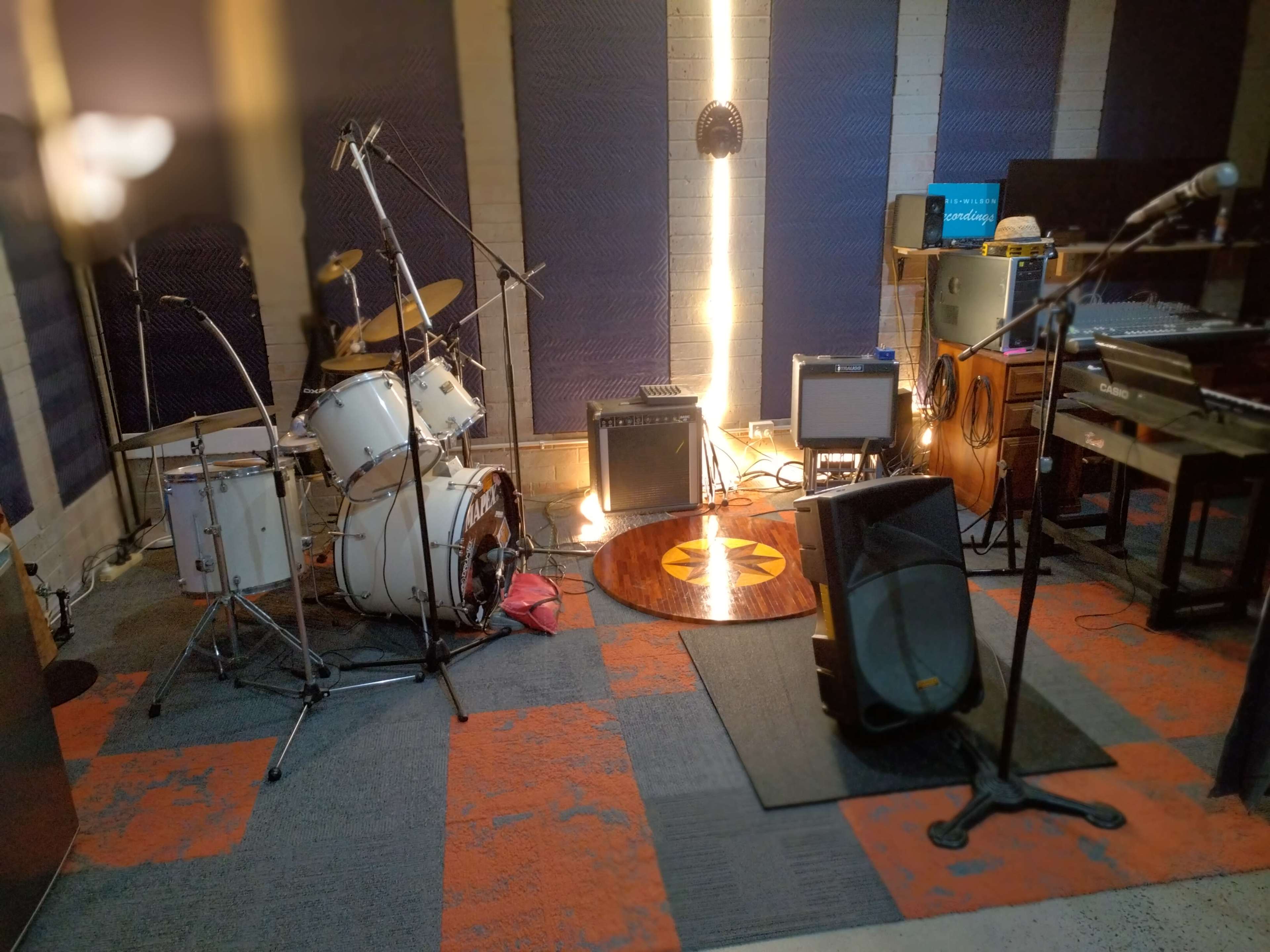 A music rehearsal studio features a drum set, amplifiers, microphones, and various instruments arranged around a central circular rug.