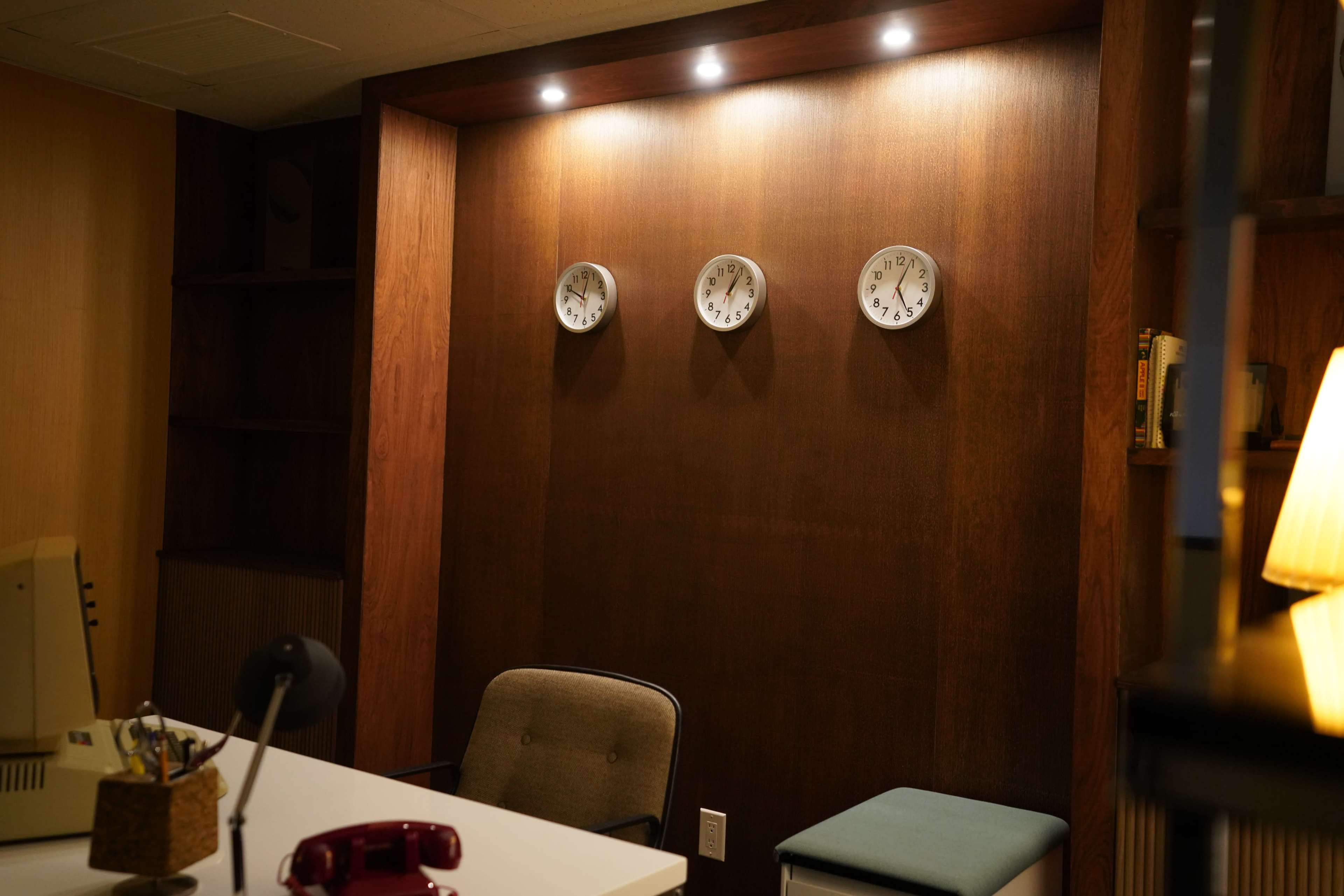 The image shows an office setting with a desk, a chair, a vintage telephone, and three wall clocks mounted on a wood-paneled wall.