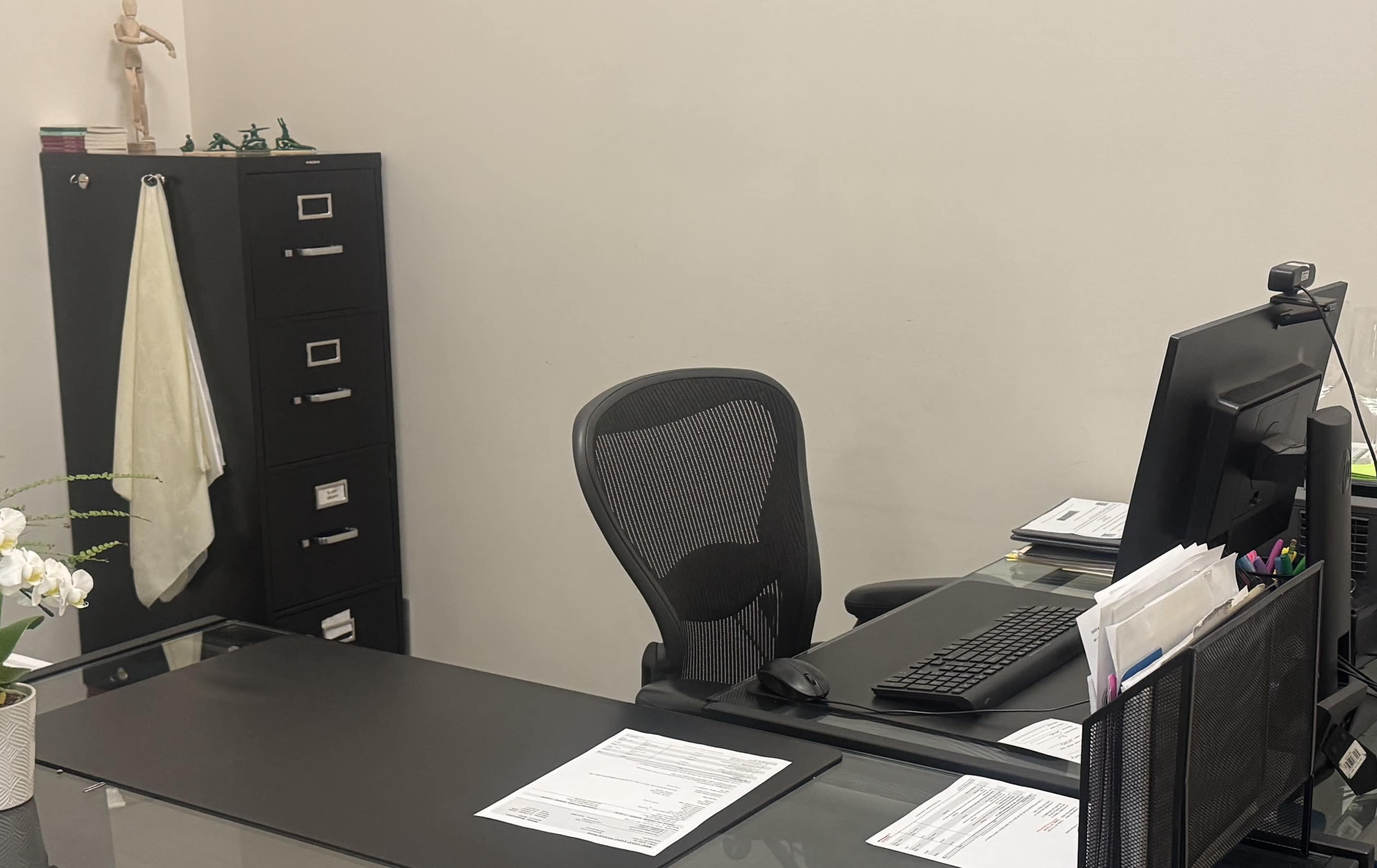 The image shows an office space featuring a black ergonomic chair, a computer monitor on a glass desk, and a filing cabinet in the background.