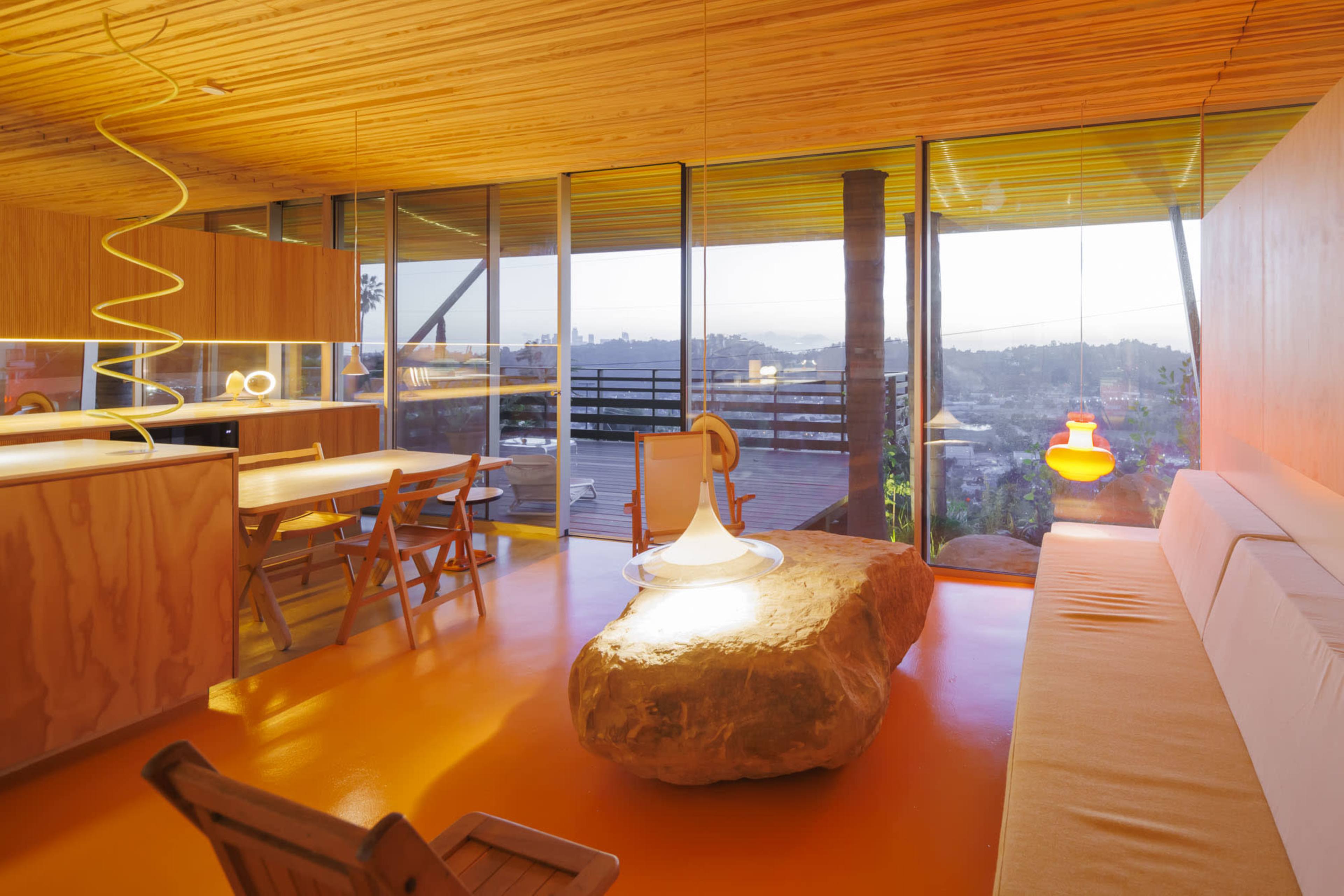 The image shows a modern interior space with large glass windows, a wooden ceiling, and minimalist furniture, featuring a central stone coffee table and colorful pendant lights.