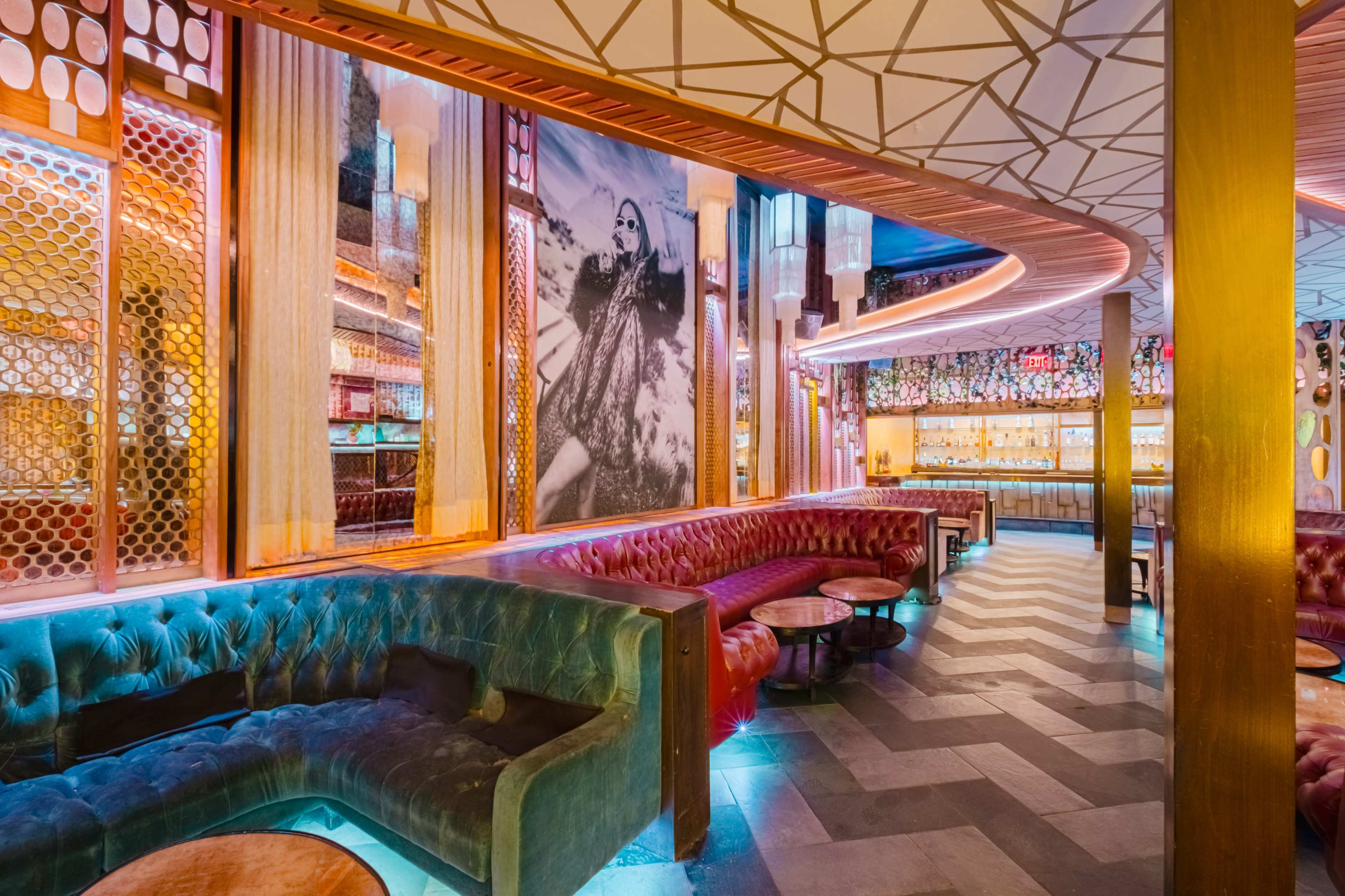 The image shows a stylish lounge with plush seating, vibrant lighting, and large wall art featuring a figure in a glamorous pose.