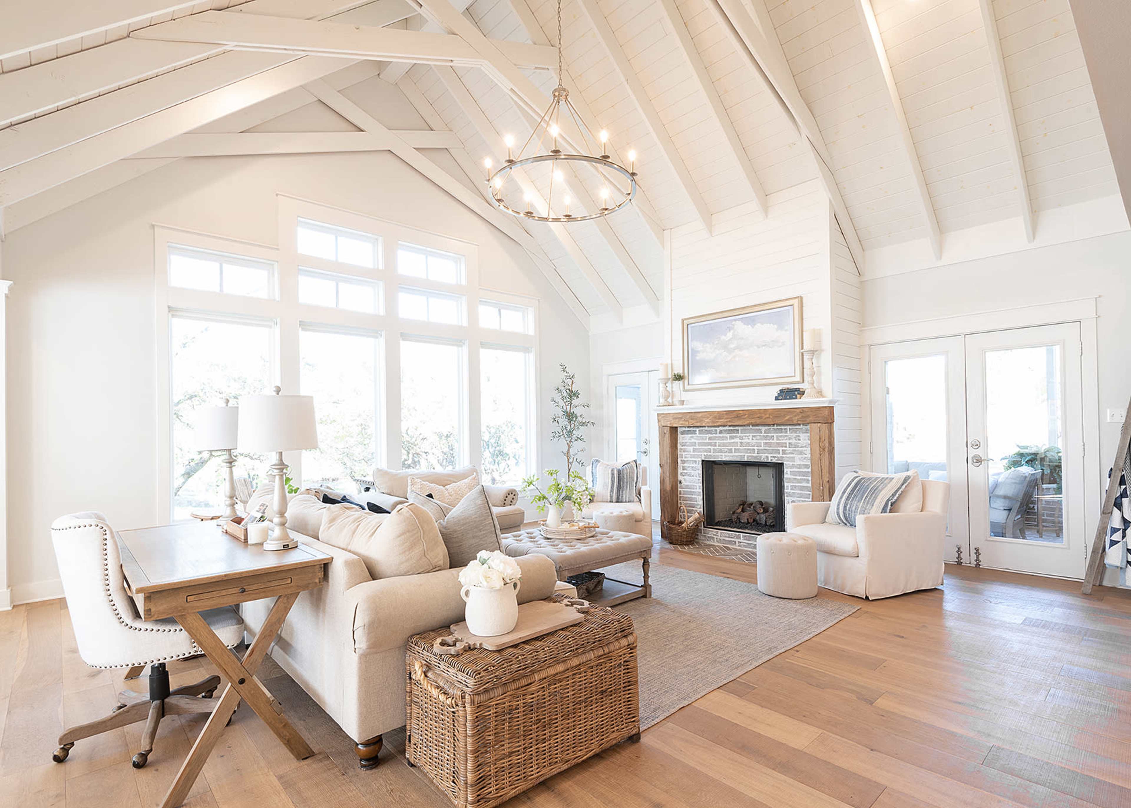 A spacious living room featuring a high vaulted ceiling, a stone fireplace, large windows, and a mix of cozy furniture arranged around a central area.