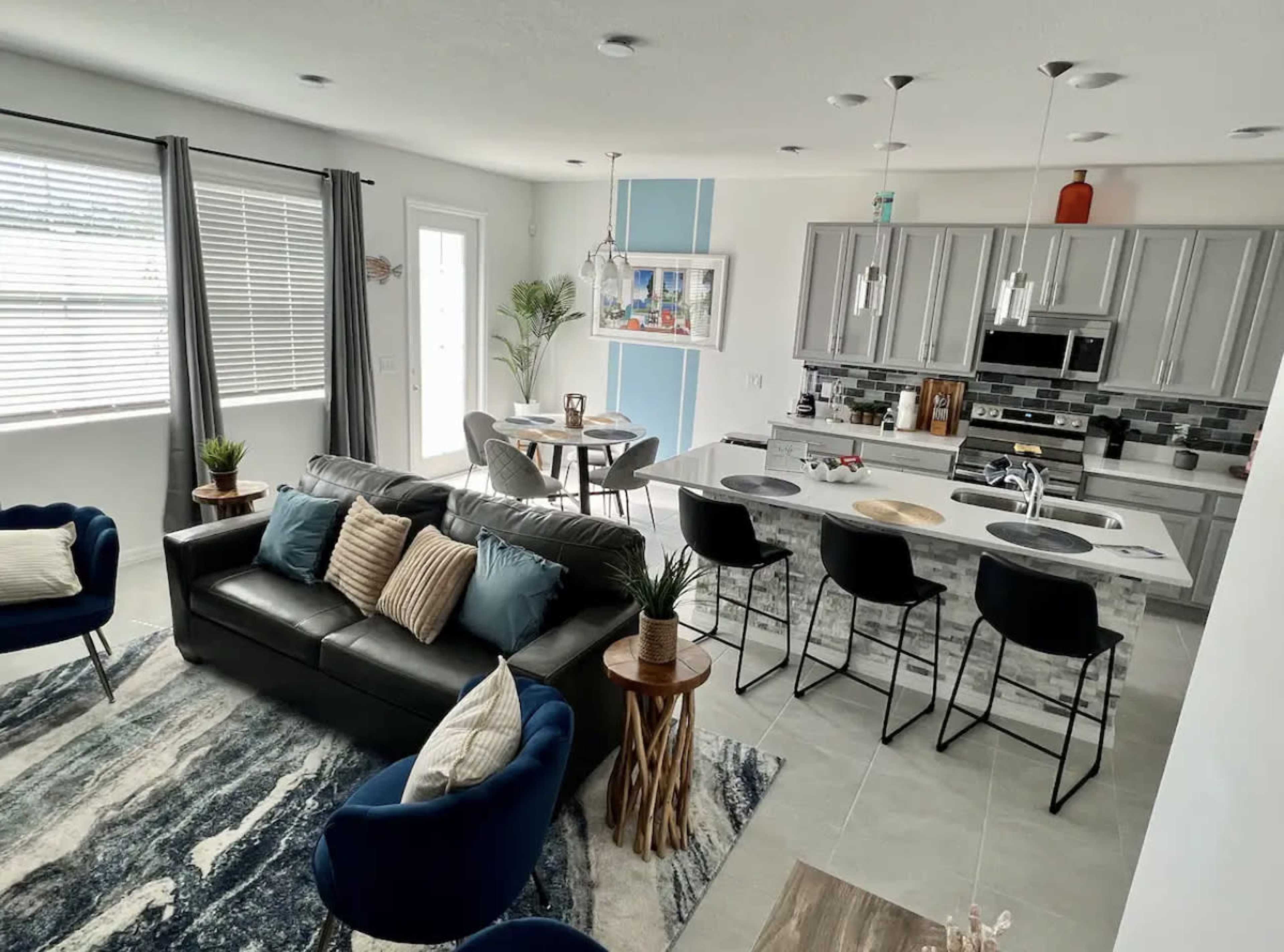 The image shows a modern living space featuring a gray sofa, blue chairs, a dining area, and a kitchen with stainless steel appliances.