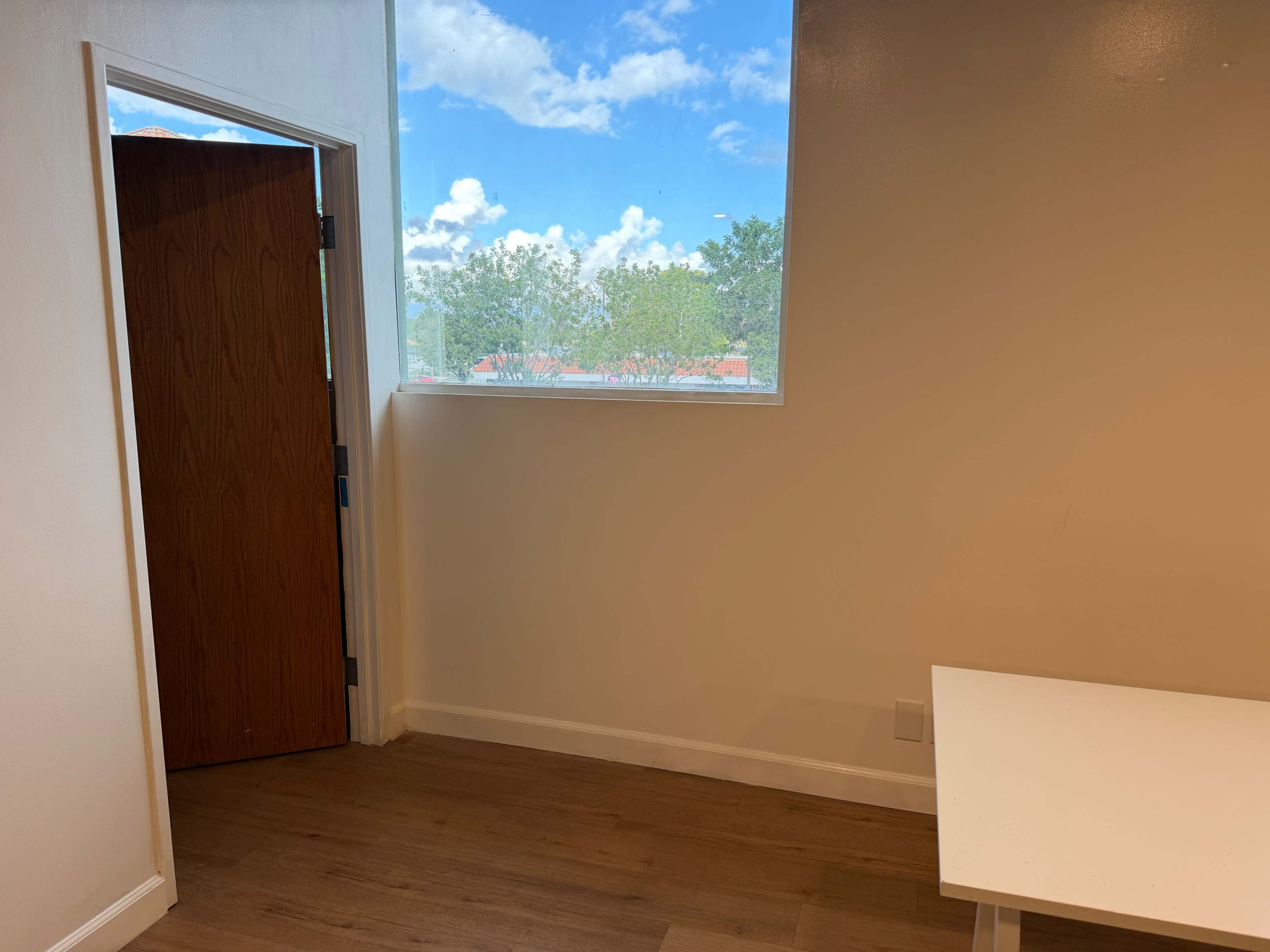 The image shows an interior room with a wooden door open, a window revealing a blue sky with clouds, and a white table against a wall.