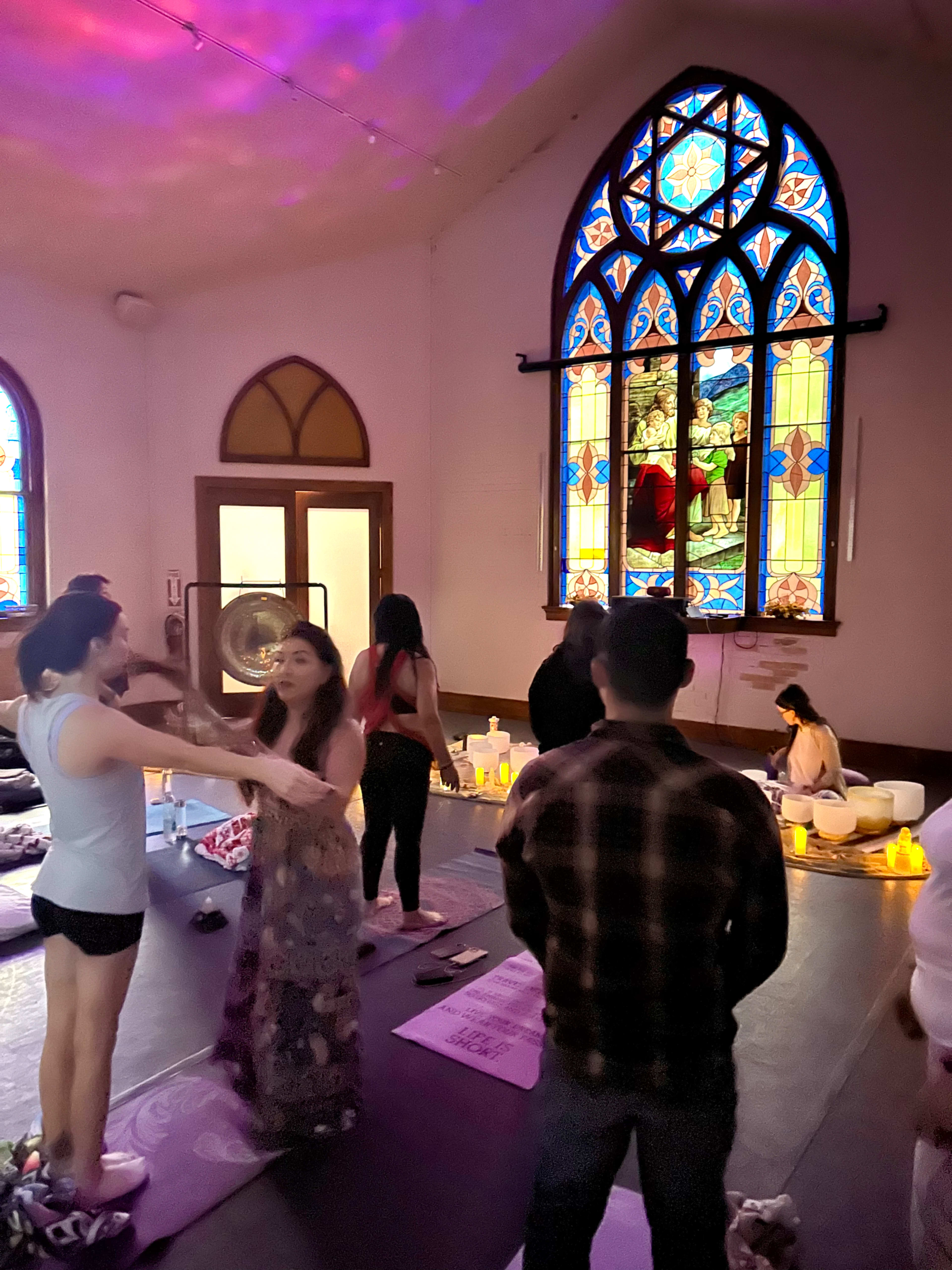 A group of people participates in a yoga session inside a room with stained glass windows depicting a religious scene.
