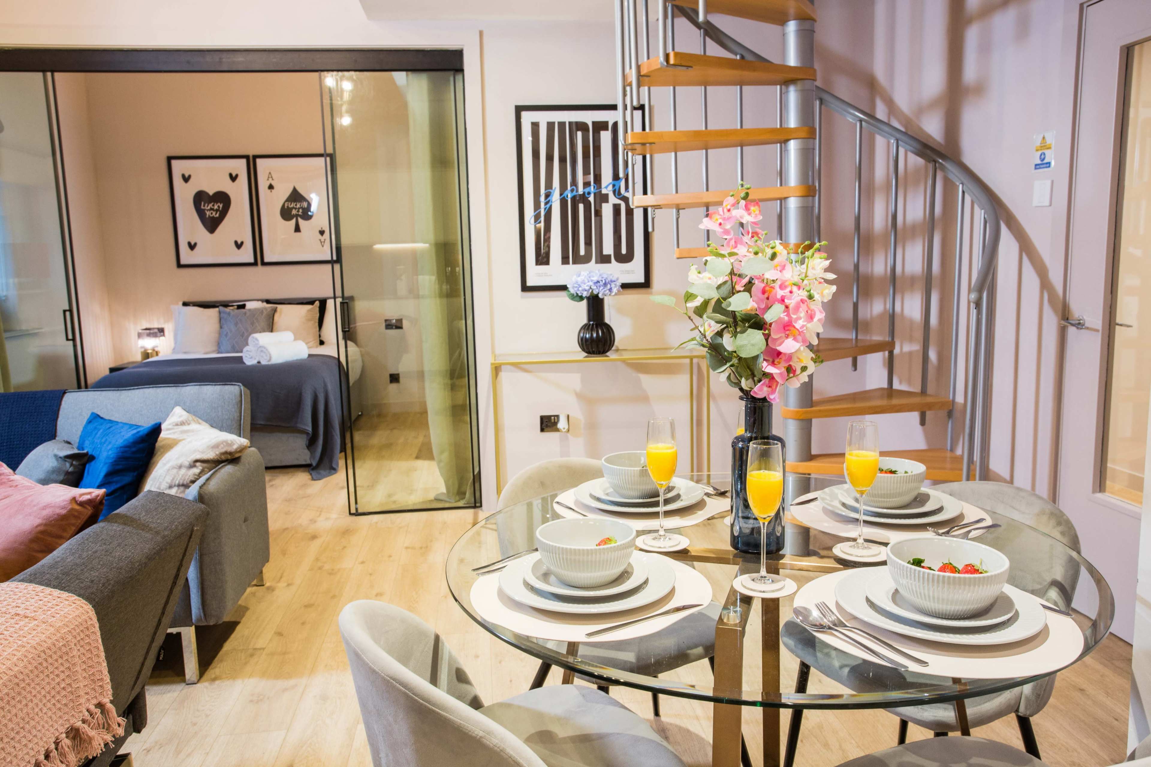 A modern studio apartment features a dining table set for four, with a spiral staircase leading to a sleeping area visible through glass walls.