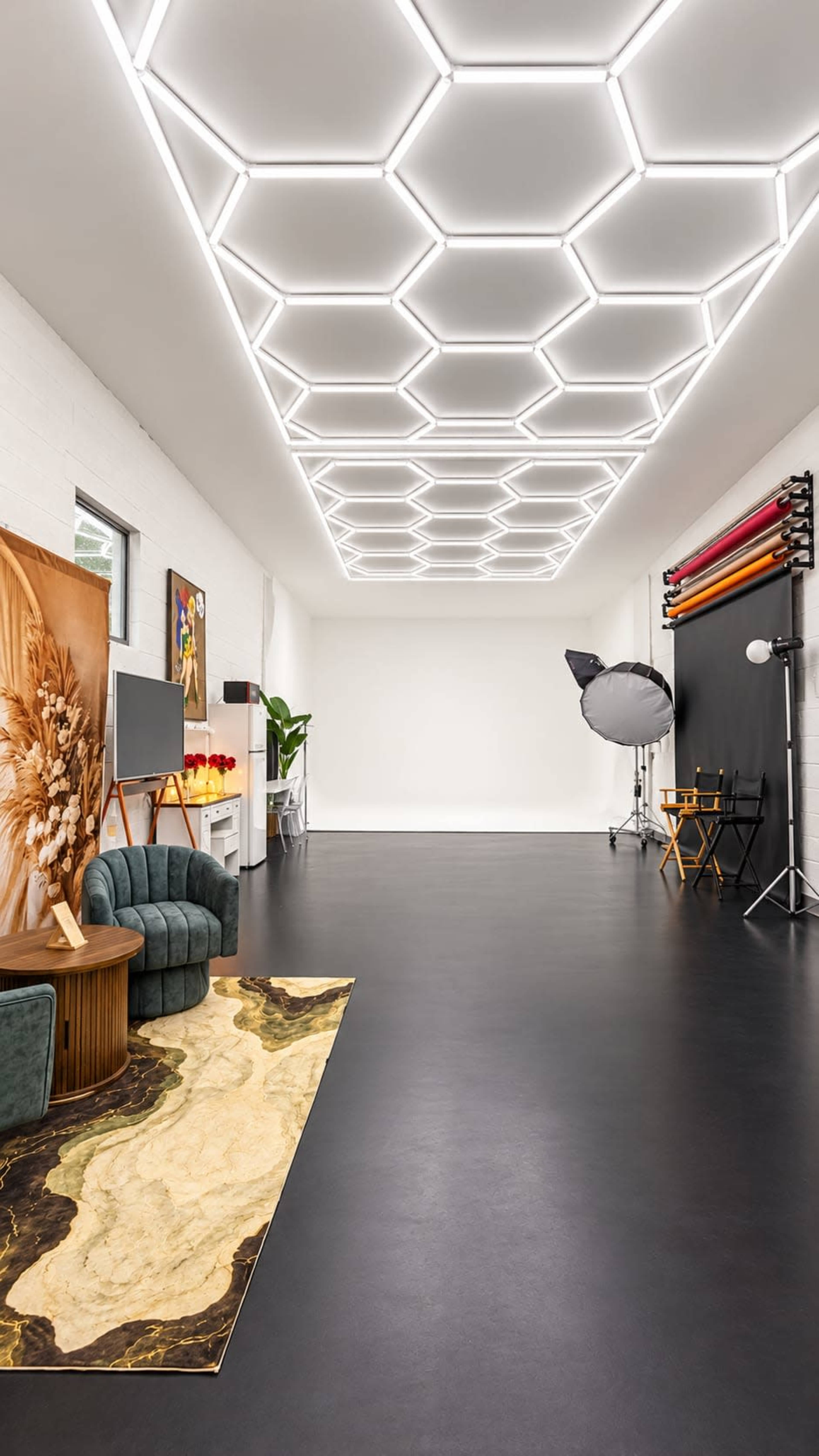 The image shows a spacious photography studio with a hexagonal light fixture ceiling, a green velvet couch, and various photography equipment set up against the walls.