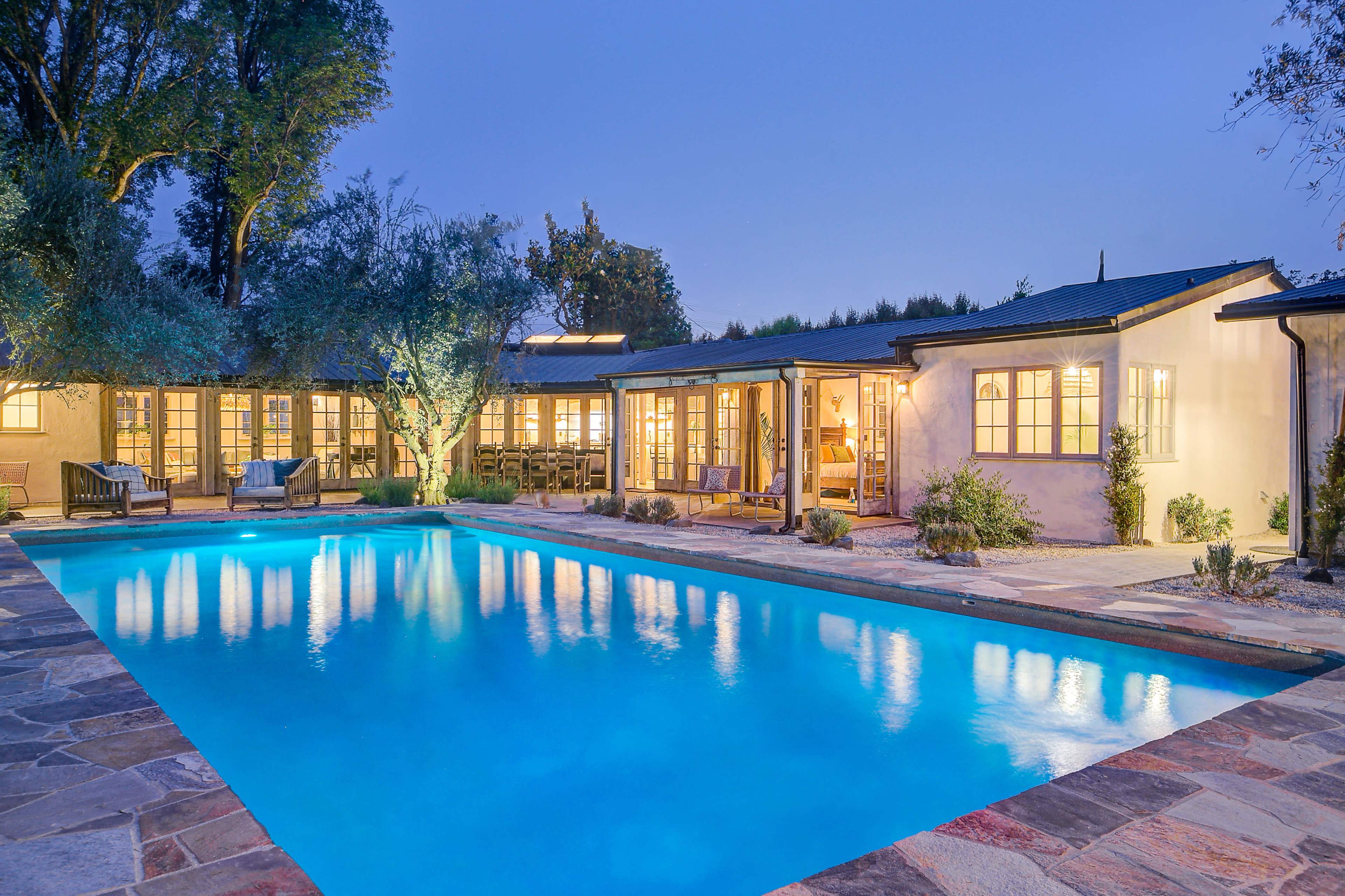 A modern house with large windows surrounded by trees, reflecting in a swimming pool illuminated at dusk.