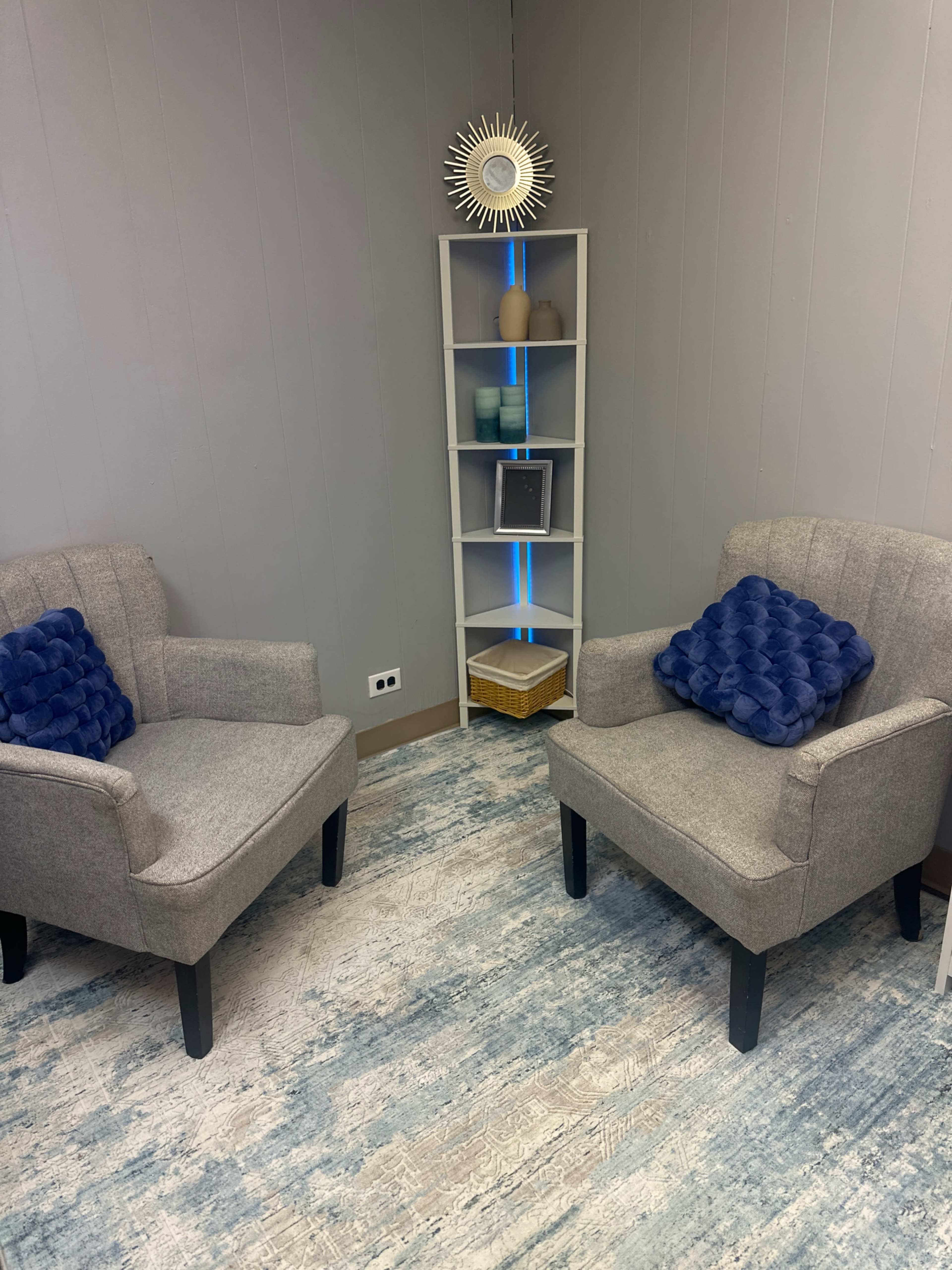 The image shows a small, cozy room with two gray armchairs facing each other, a shelf with decorative items, and a blue-lit accent against the wall.