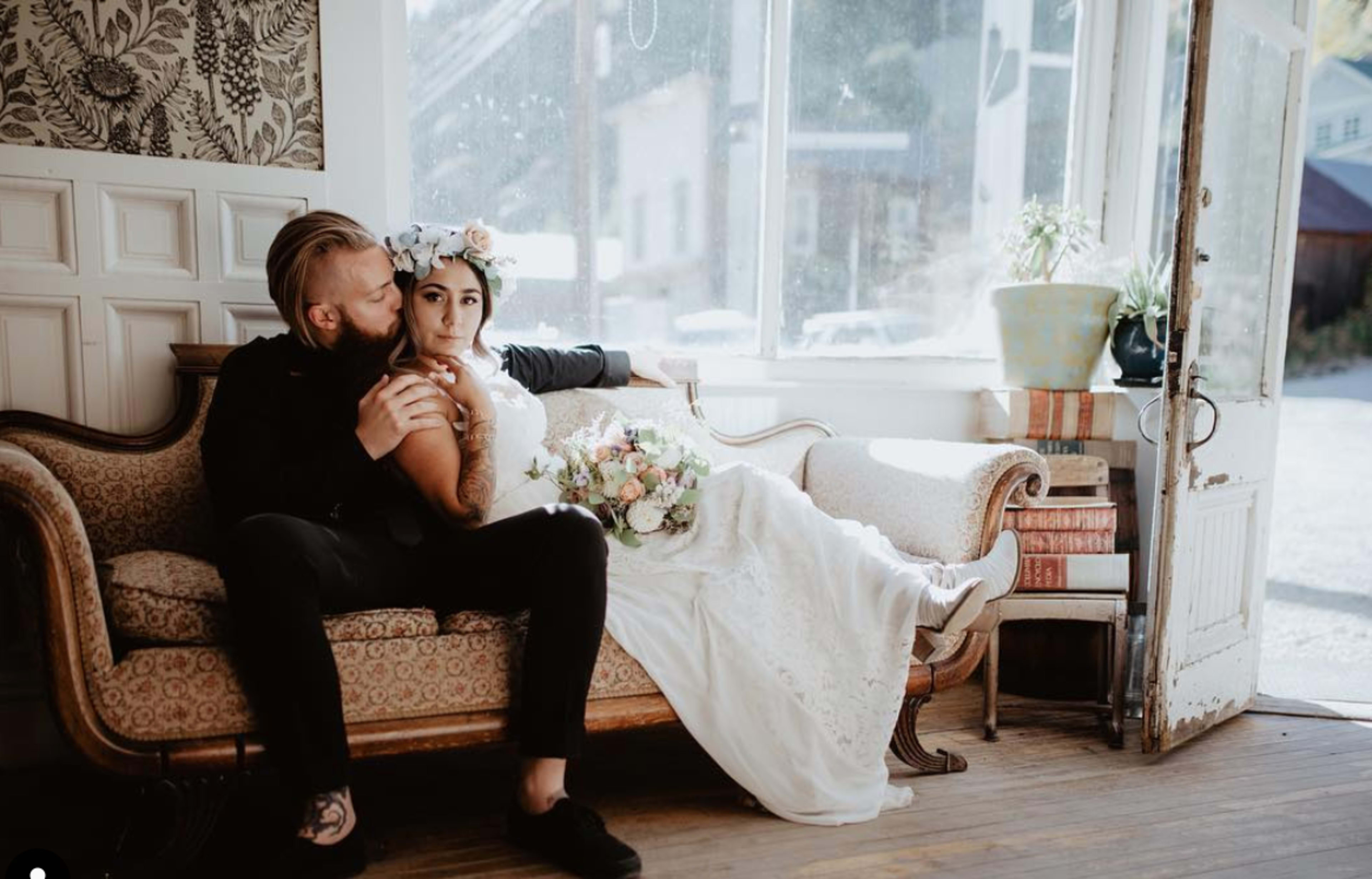 A couple sits together on a vintage couch in a sunlit room, with the bride holding a floral bouquet.