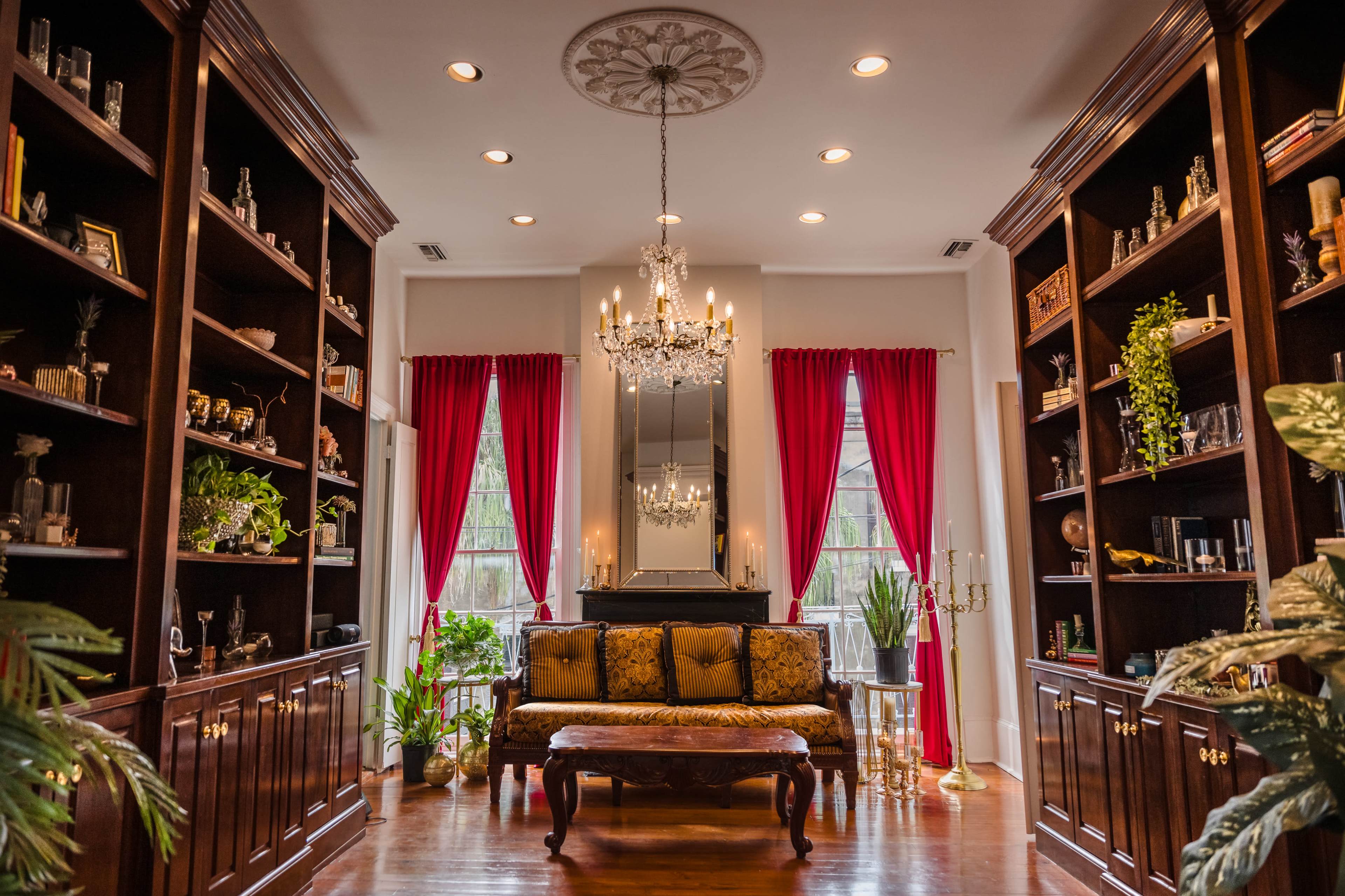 The image shows an elegantly decorated living room with dark wooden shelves, a central chandelier, and a plush sofa surrounded by plants and framed photos.