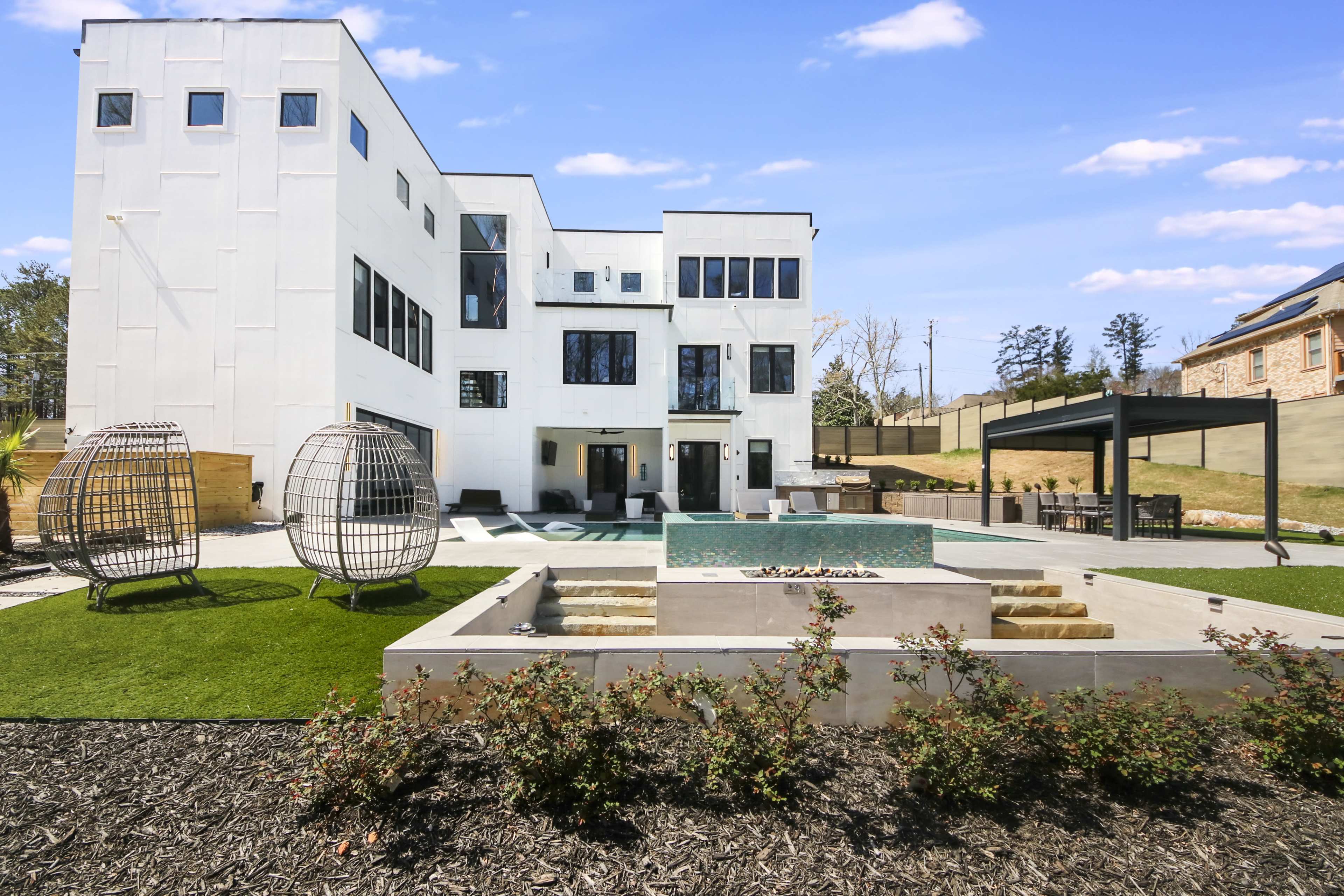 A modern white house with large windows, a swimming pool, and a landscaped backyard featuring seating areas and a covered gazebo.