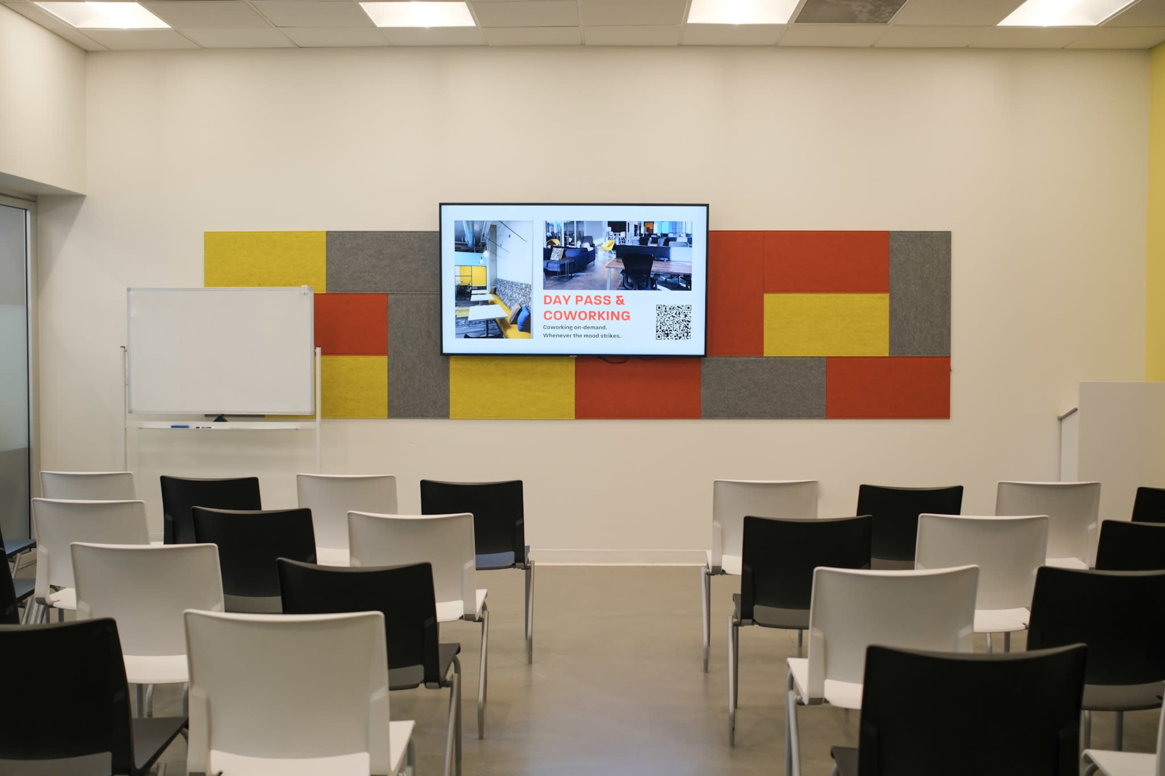 A modern coworking space features a screen displaying information about day passes, with rows of empty white chairs arranged in front of it.