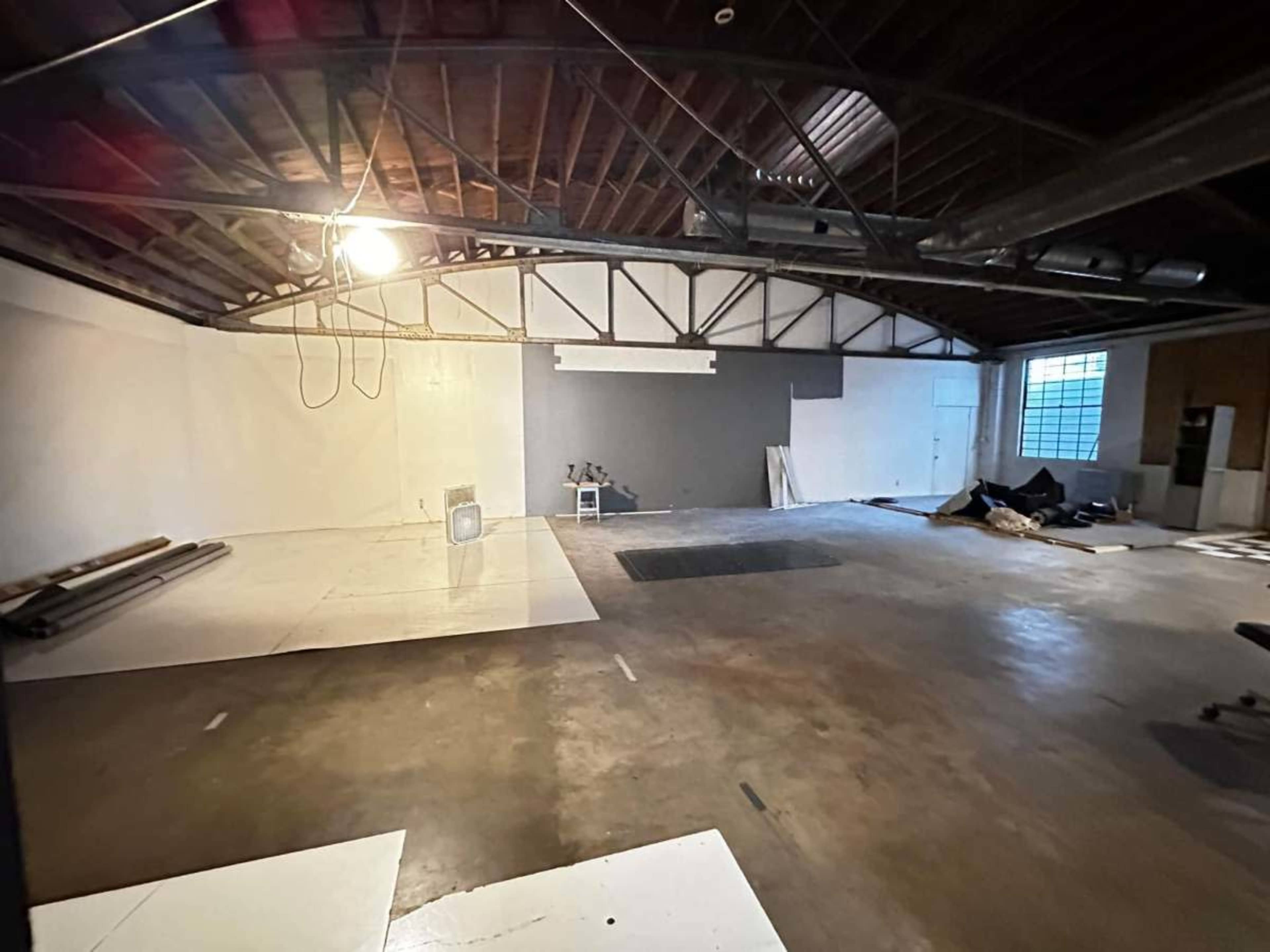 The image shows an empty industrial space with unfinished flooring, exposed beams, and a few scattered items against the walls.