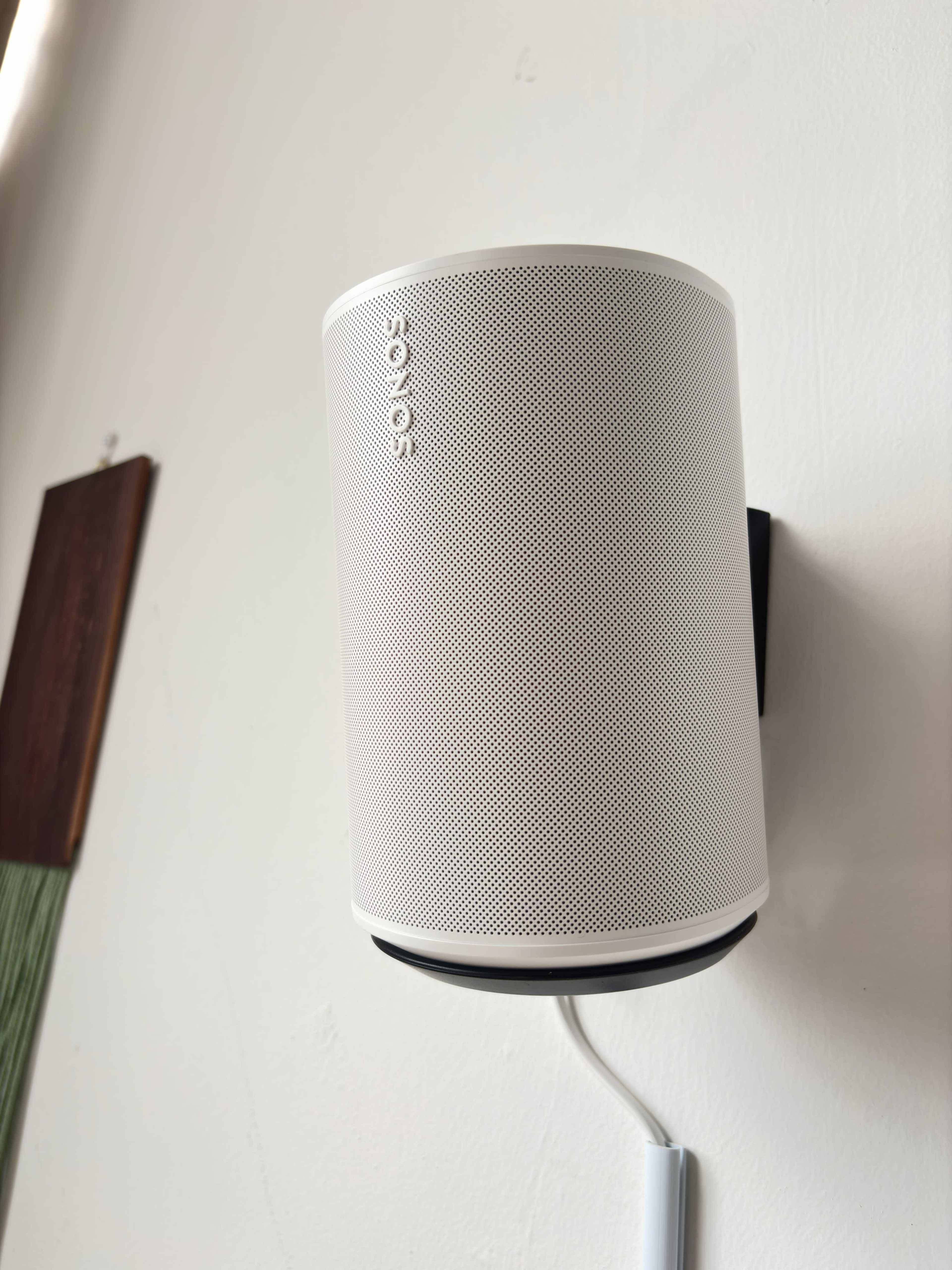 A white Sonos speaker is mounted on a wall with a cable running down to the floor.
