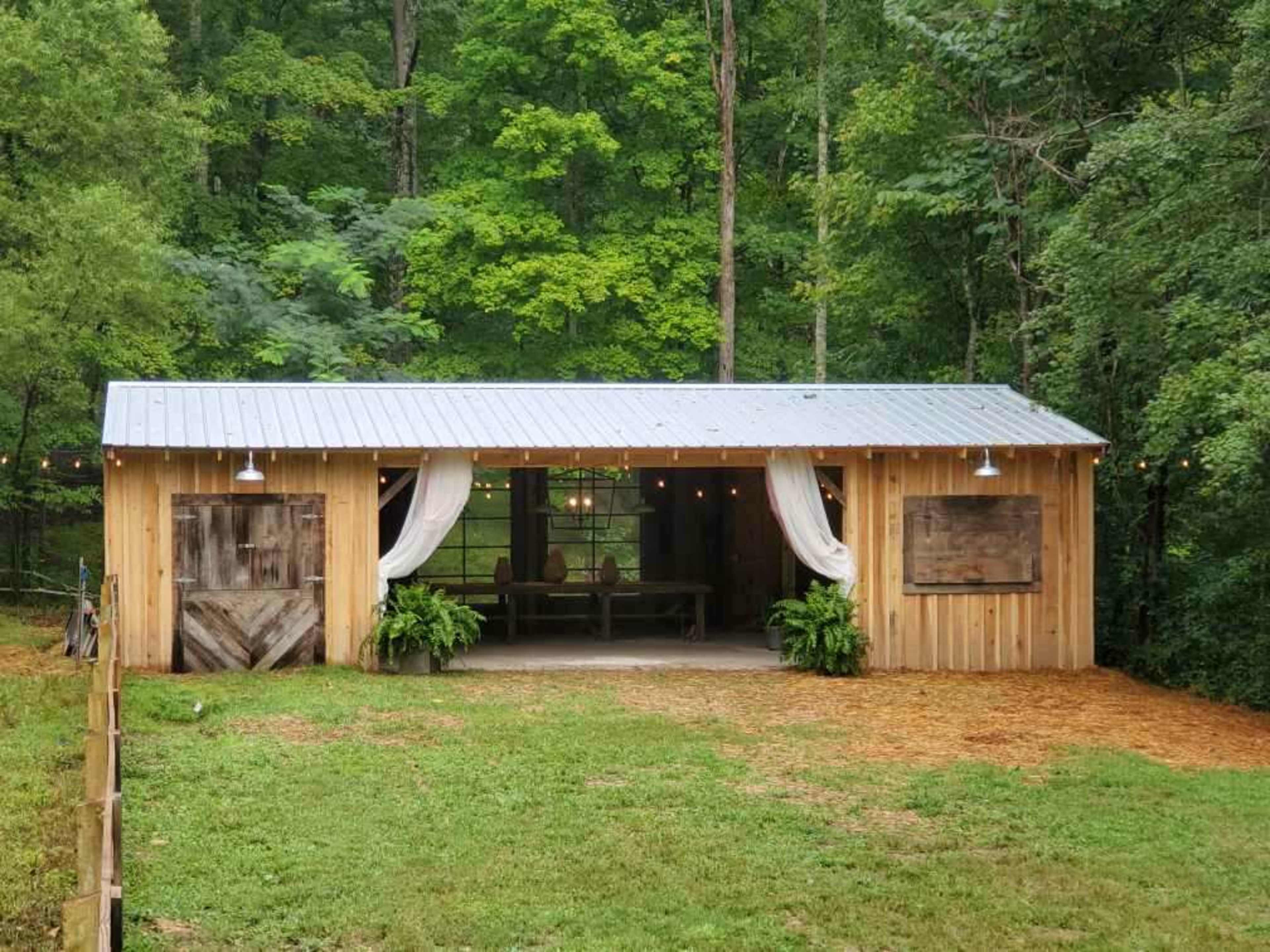 A wooden cabin with a metal roof features open sides, decorative curtains, and is surrounded by greenery.