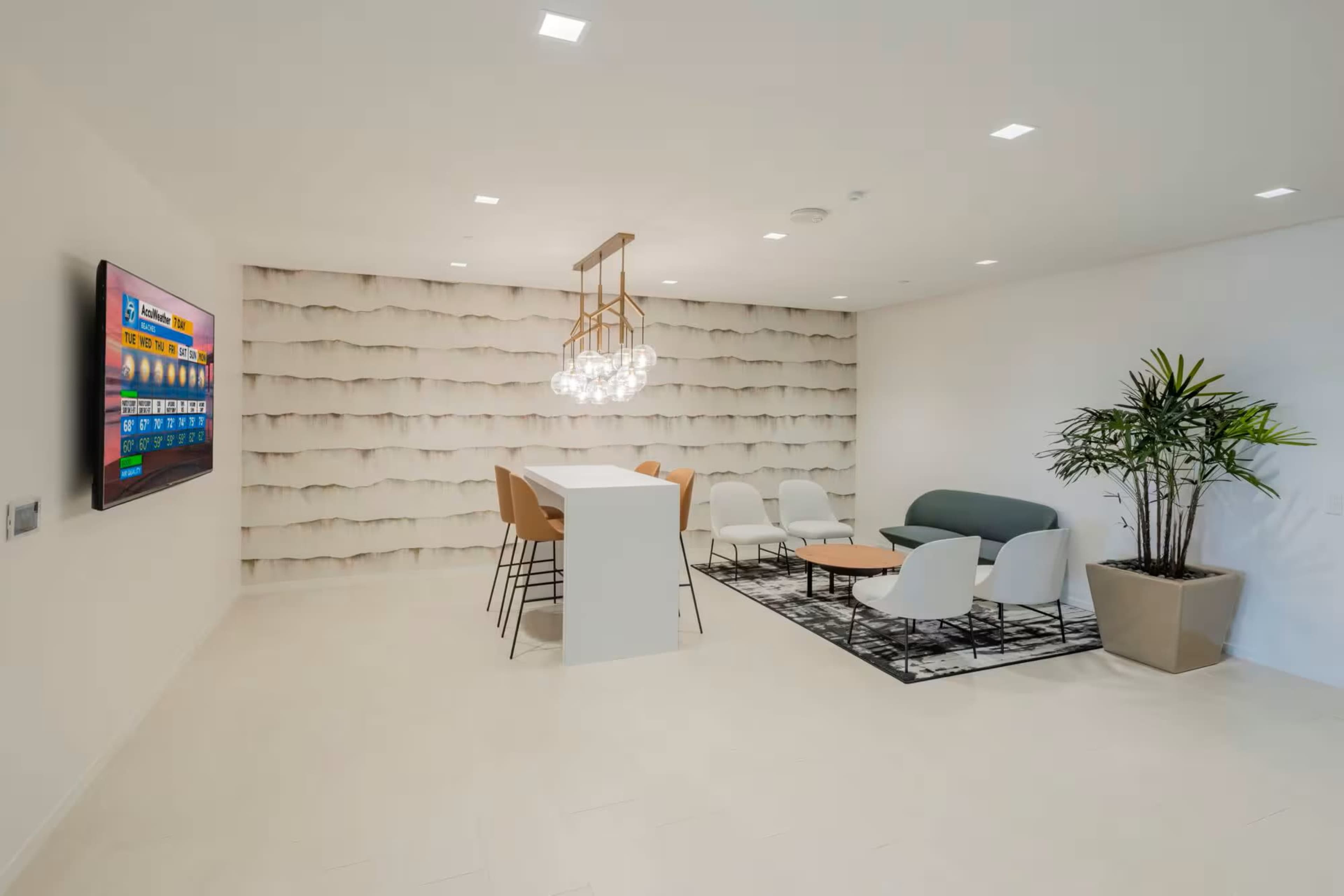 The image shows a modern lounge area featuring a light-colored wall with textured design, a dining table with high chairs, a sofa, white chairs, and a large TV mounted on the wall.