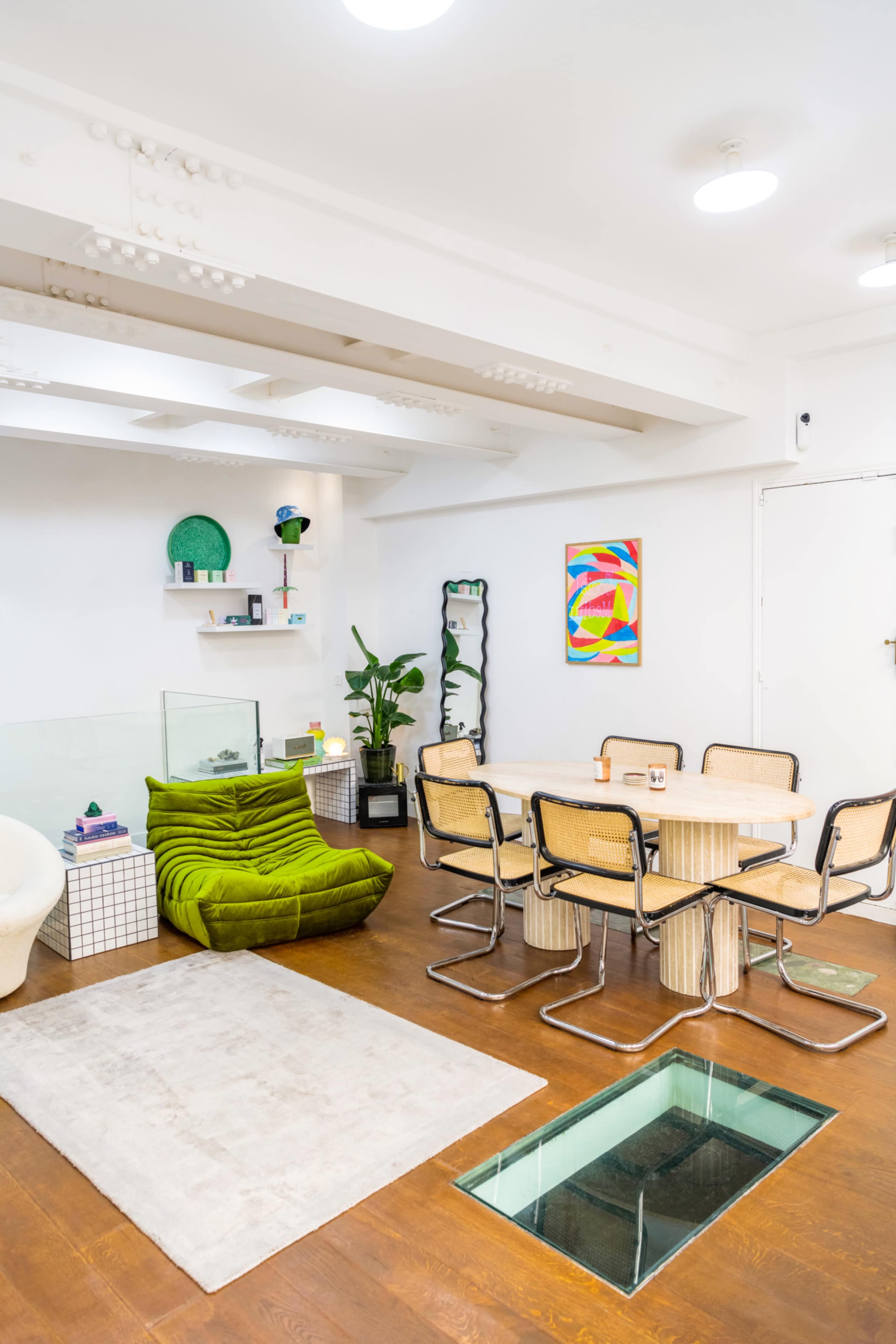 A modern interior space with a green chair, a round table surrounded by metal chairs, and a glass floor section, featuring decorative items and artwork on the walls.