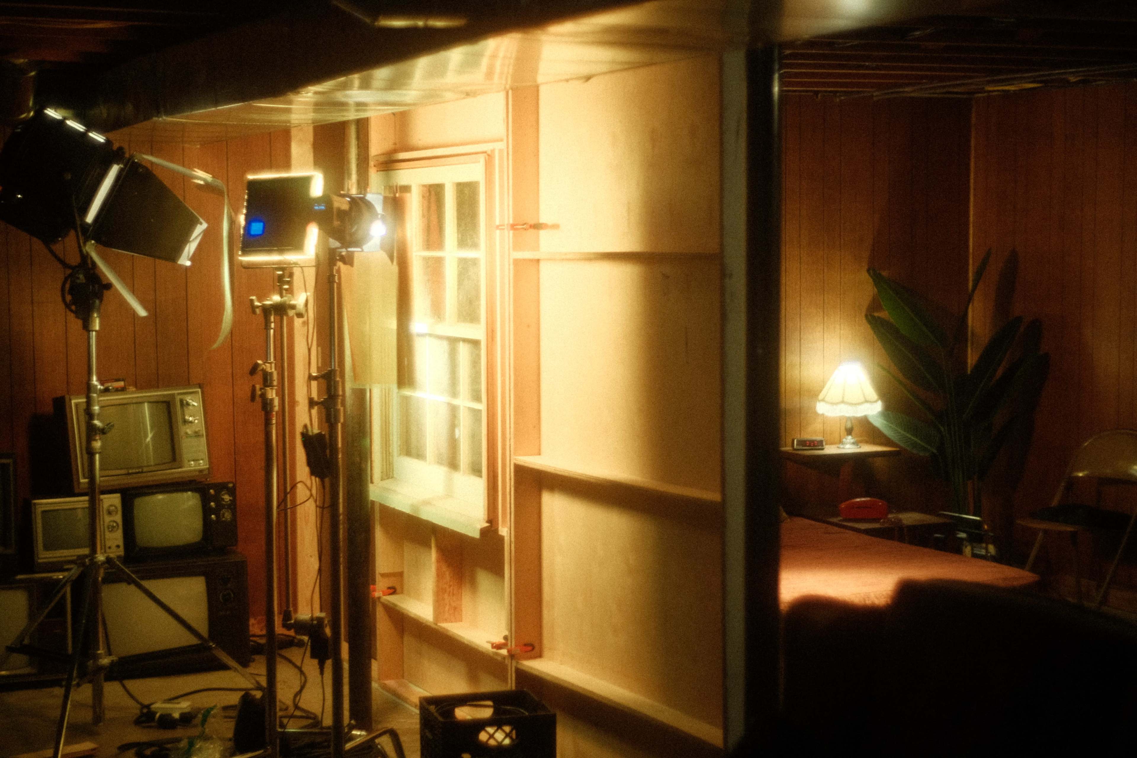The image shows a dimly lit basement scene with film equipment, including lights and monitors, alongside a window, a plant, and a lamp on a small table.