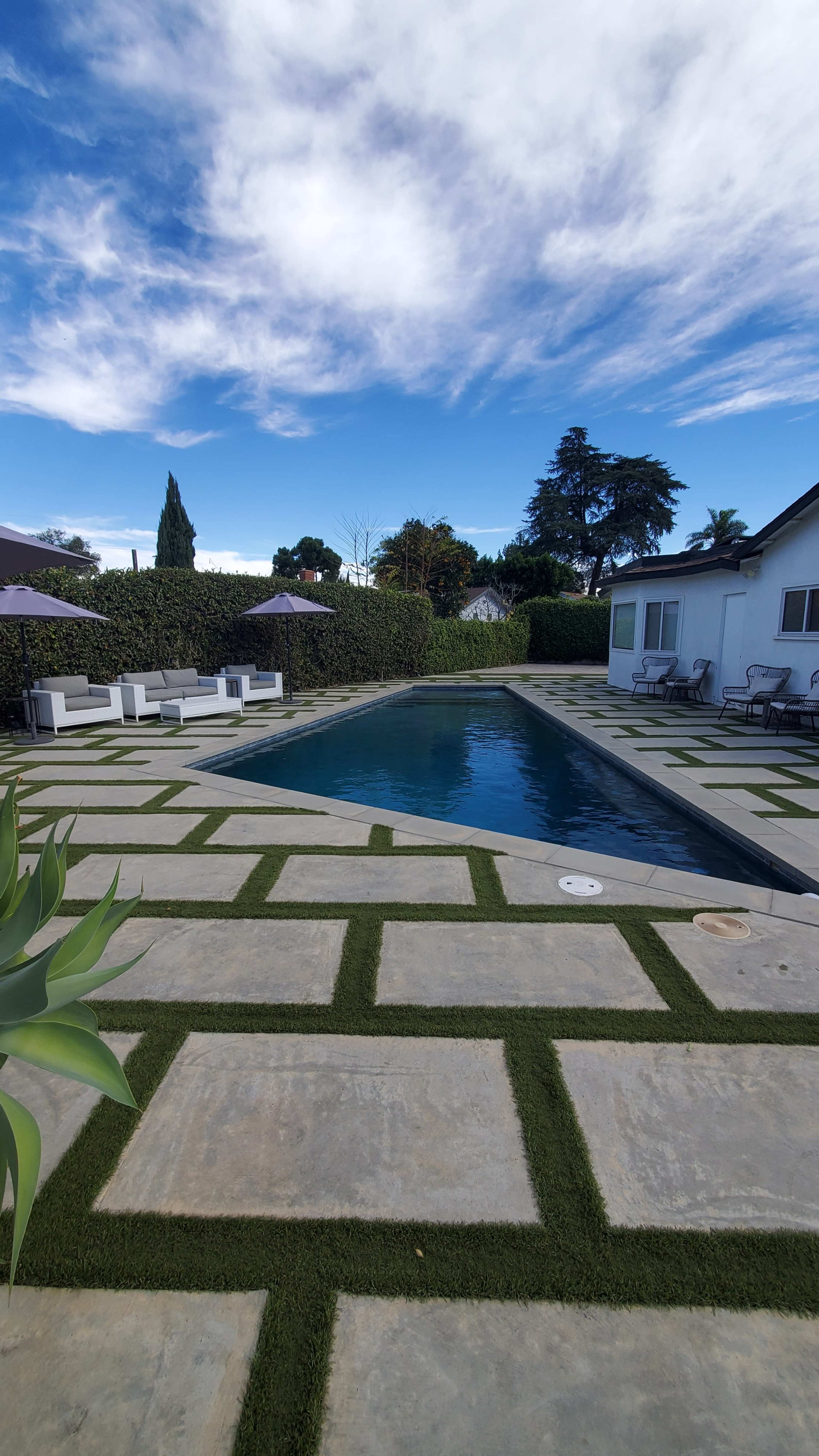 The image shows a rectangular swimming pool surrounded by a patterned concrete patio with sections of grass, under a blue sky with scattered clouds.