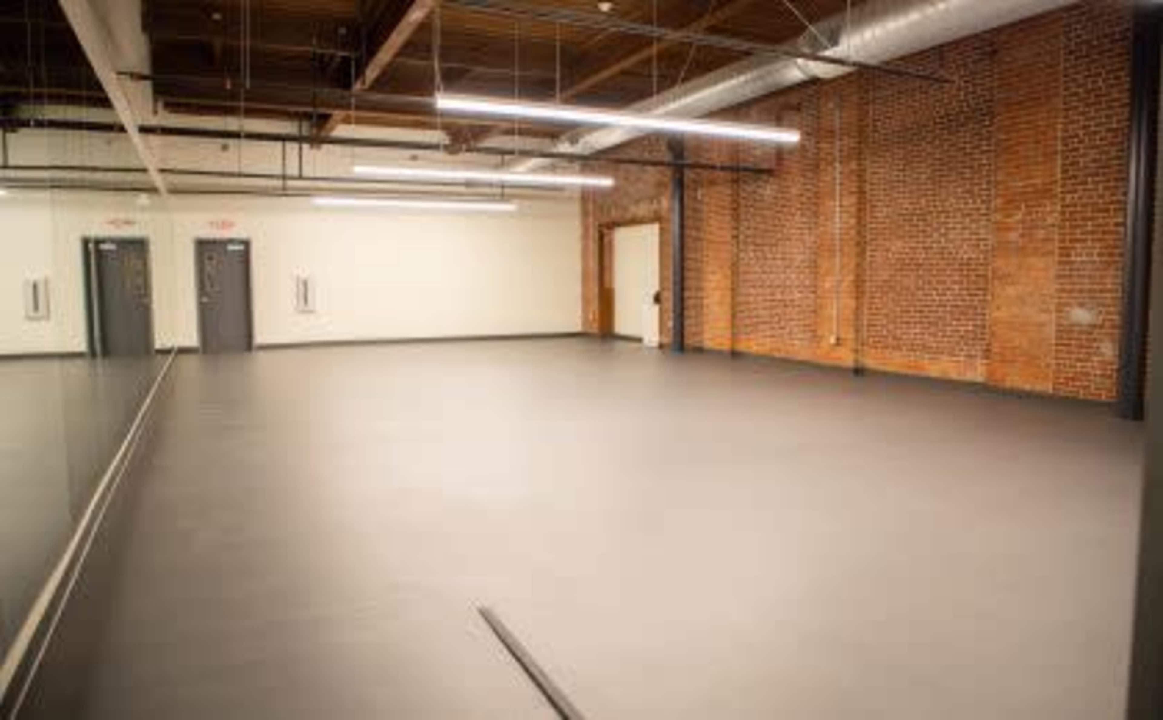 A spacious, empty room with a smooth gray floor, exposed brick walls, and overhead lighting.