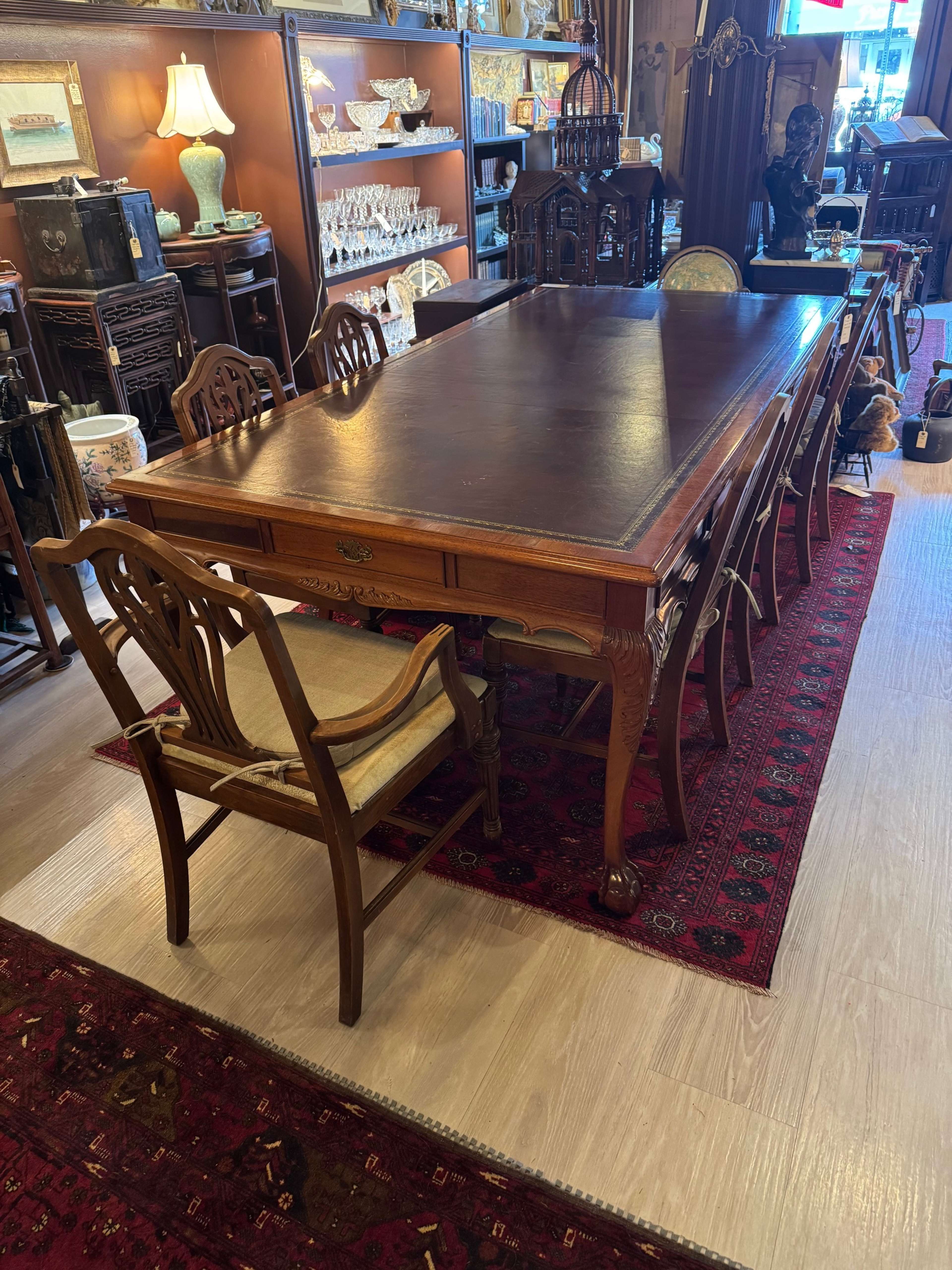 The image shows a large wooden dining table surrounded by chairs in an antique shop, with various decorative items displayed in the background.