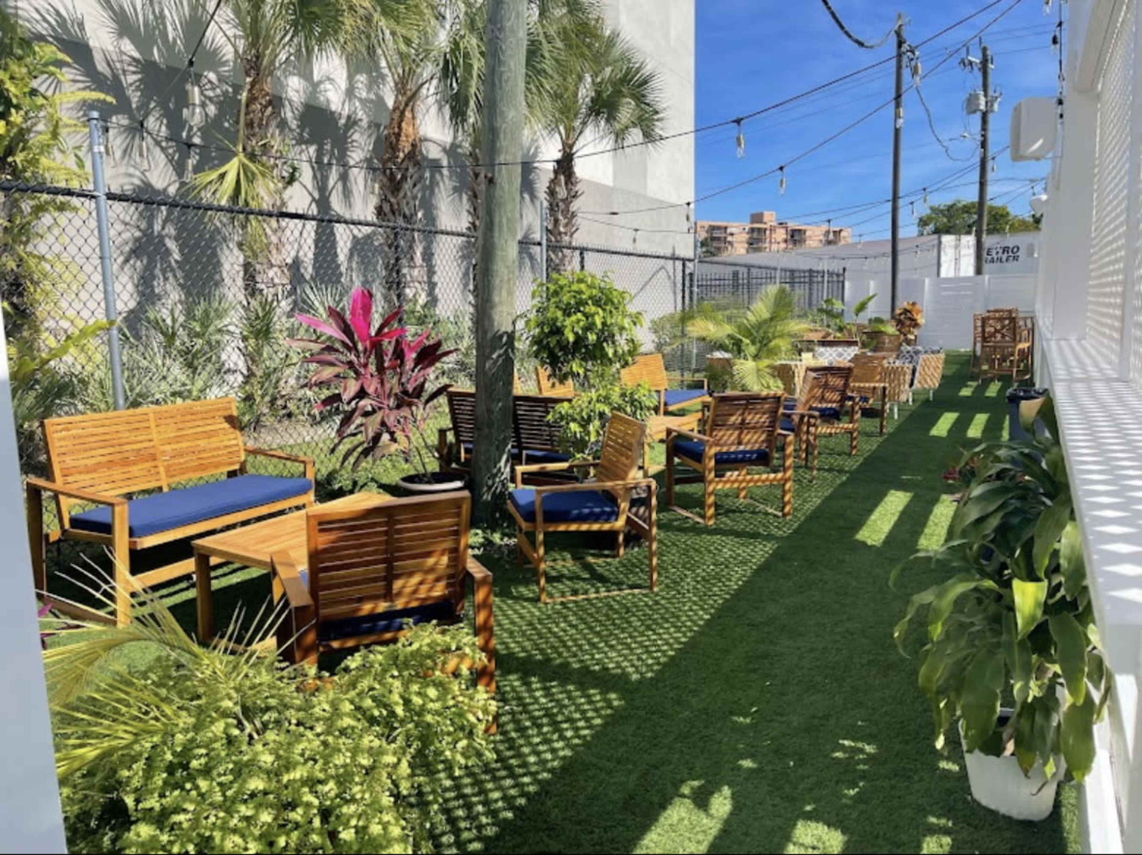 A landscaped outdoor seating area with wooden furniture, potted plants, and artificial grass, enclosed by a fence and palm trees.