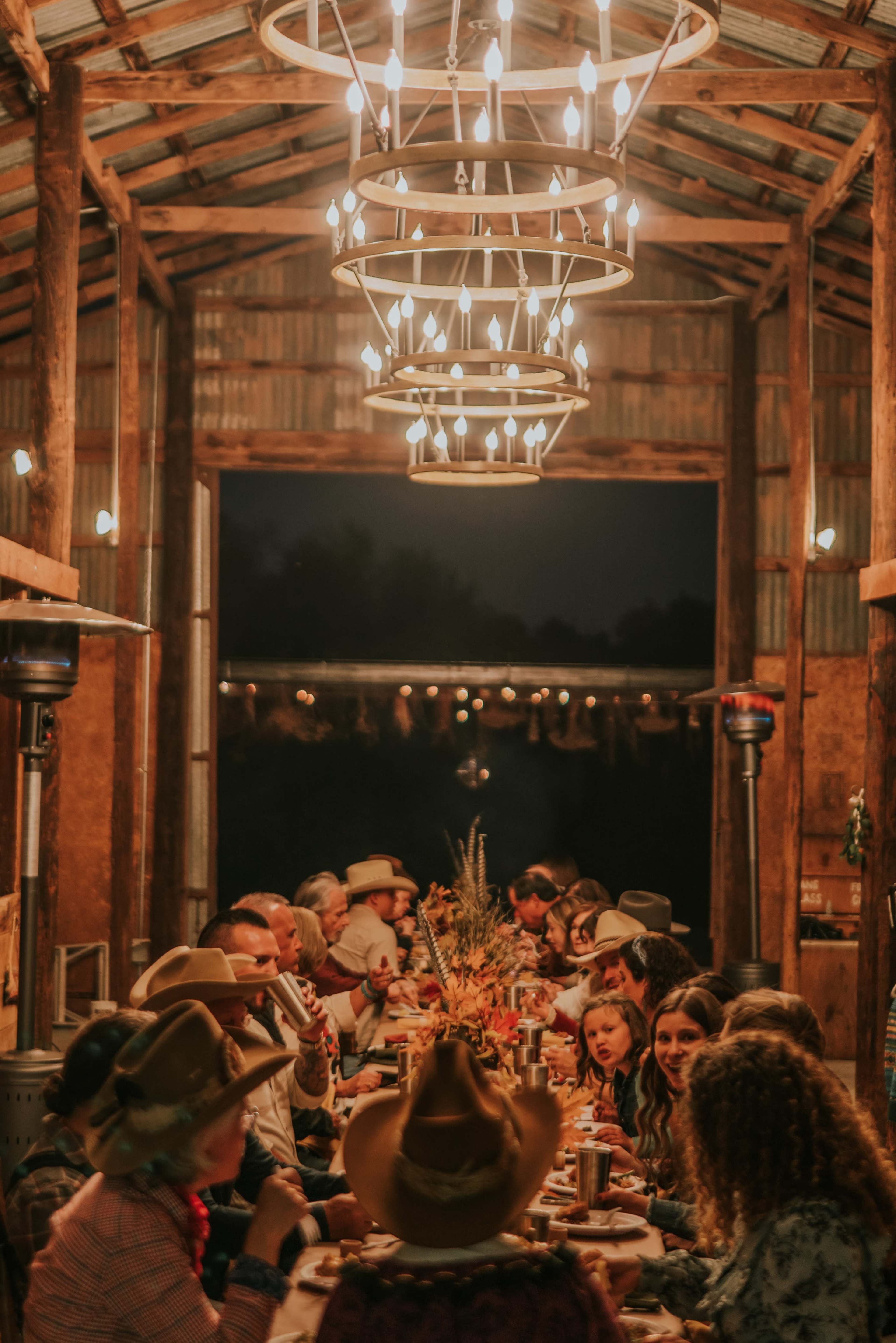 A long table set for a dinner gathering is illuminated by large circular chandeliers in a rustic barn at night.