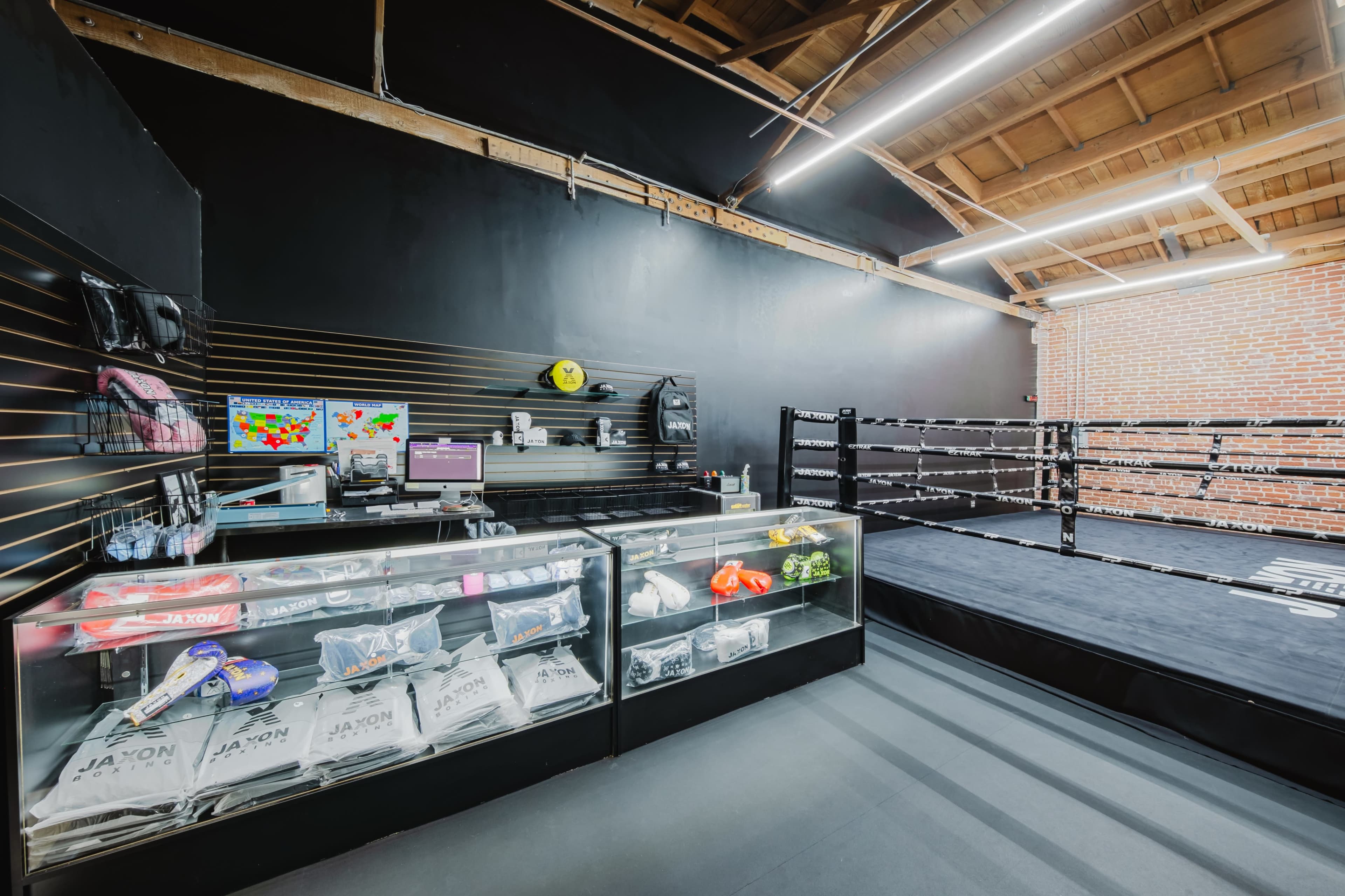 The image shows a boxing gym interior featuring a training ring, display shelves with equipment, and a glass counter.