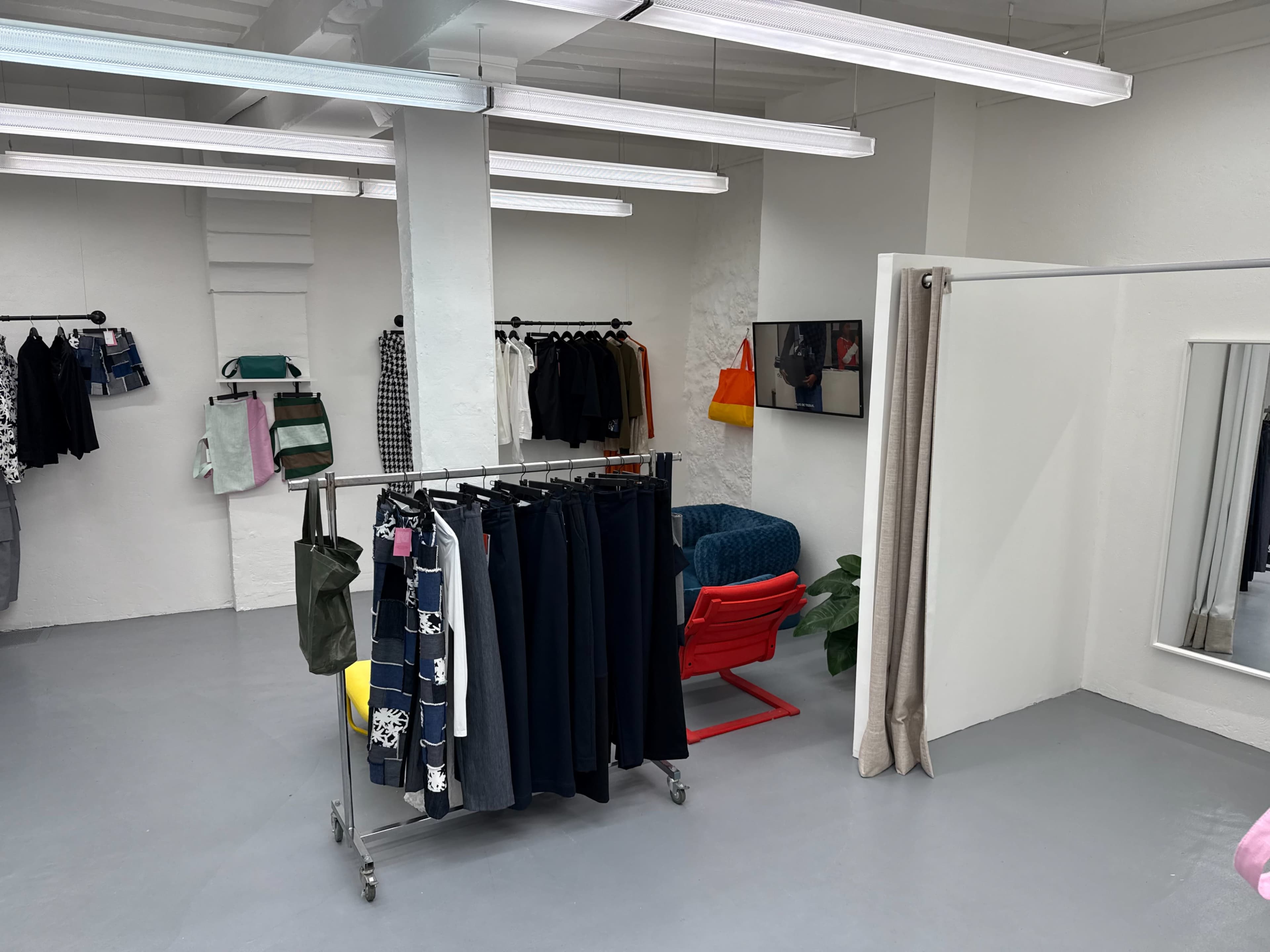 The image shows a minimalist retail space featuring clothing racks, a seating area, and a large mirror against a white wall.