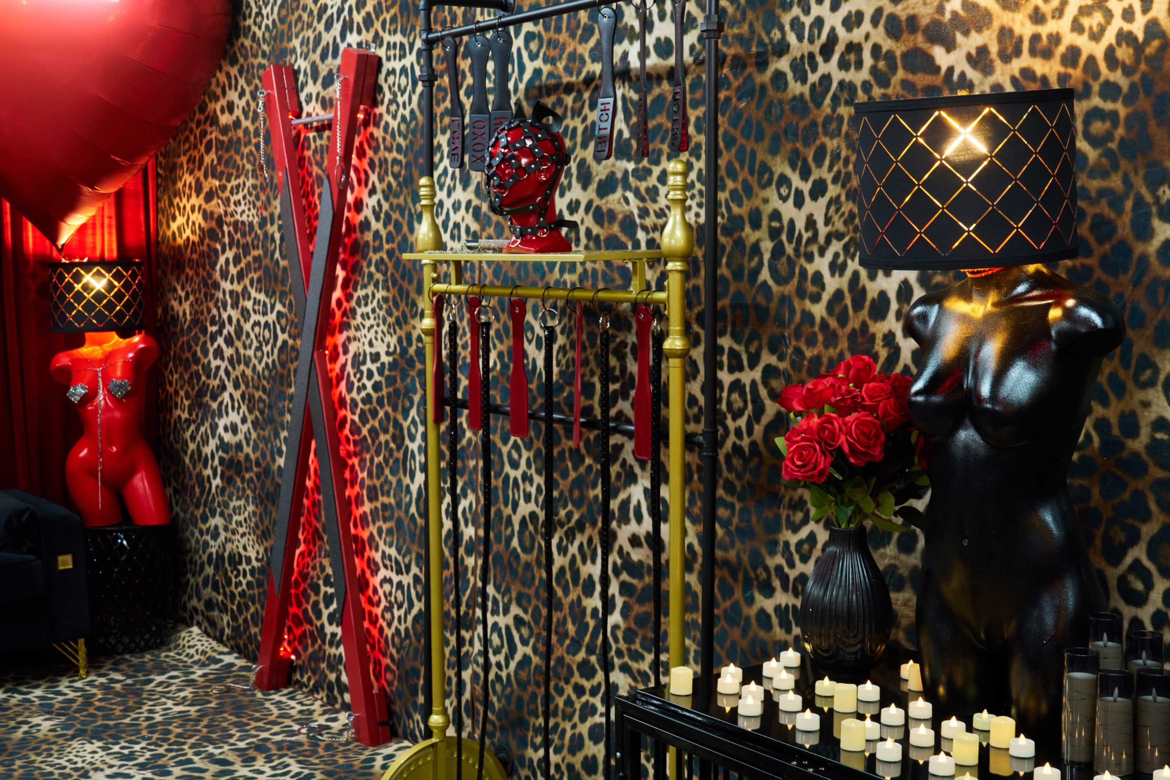 The scene features a room decorated with leopard print wallpaper, displaying red and black mannequins, roses, and various items related to bondage and decor.