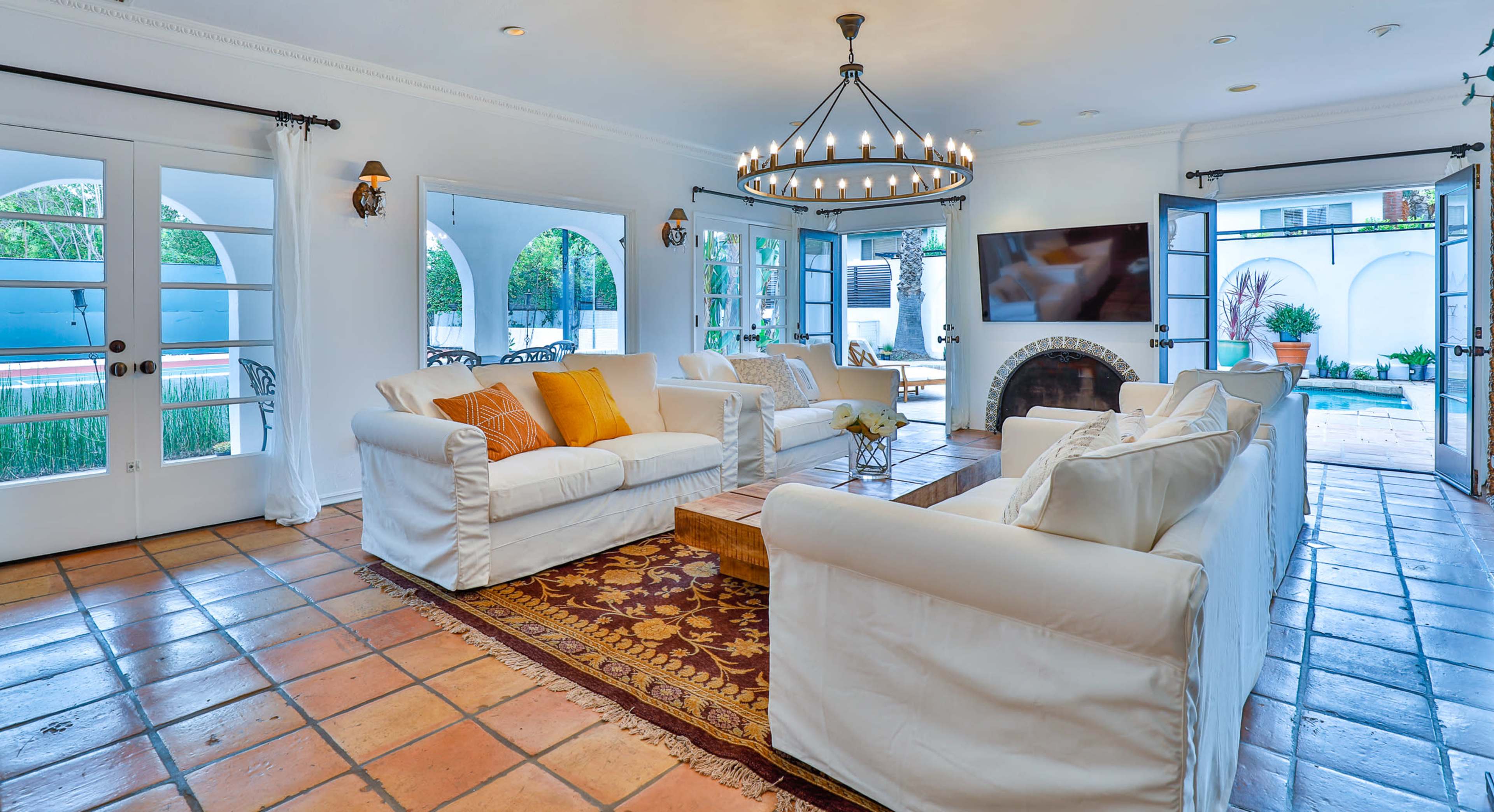 A bright living room with white sofas, a wooden coffee table, and a large chandelier, framed by arched doorways leading to a patio.