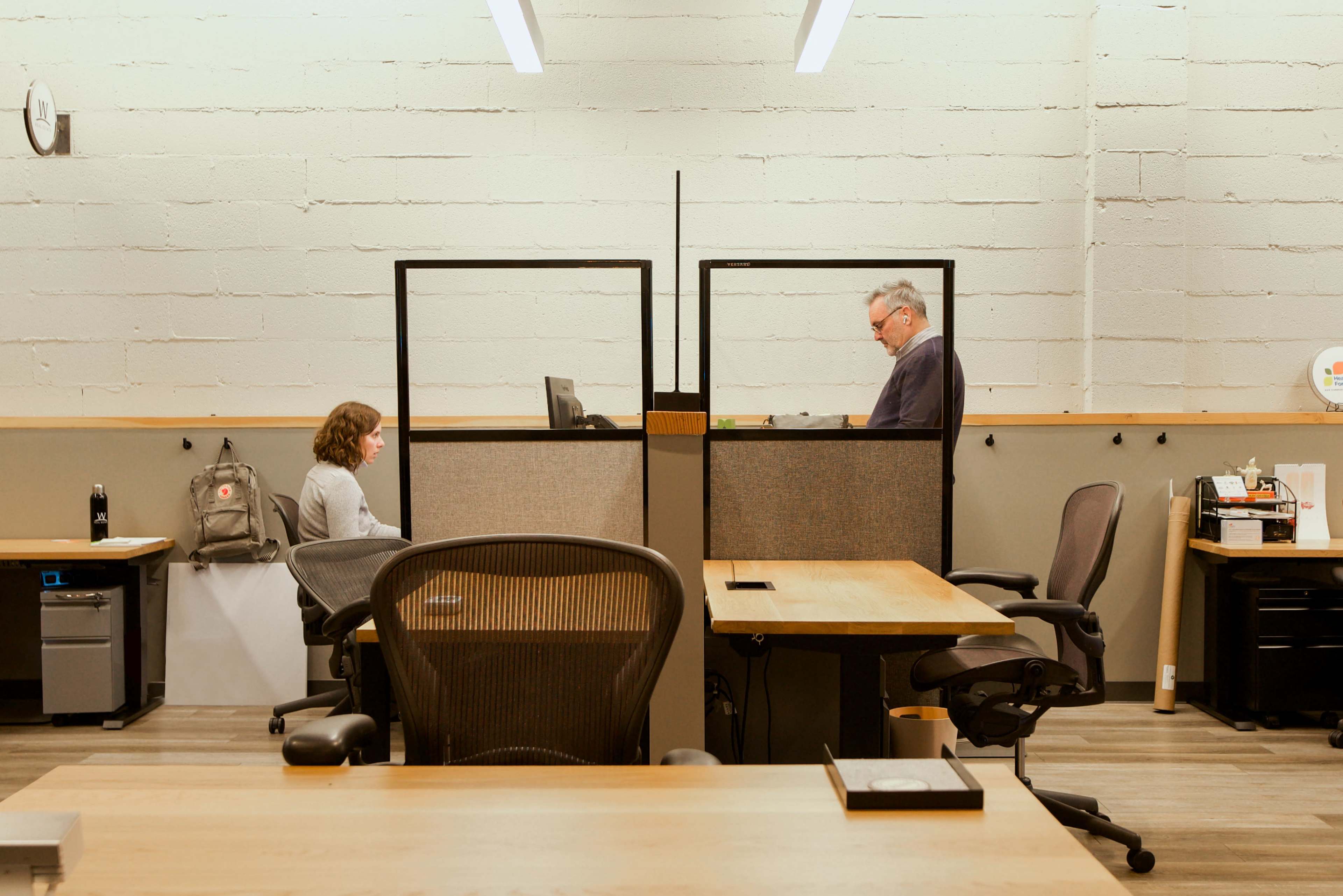 Two people work at separate desks in an office space divided by low partitions.