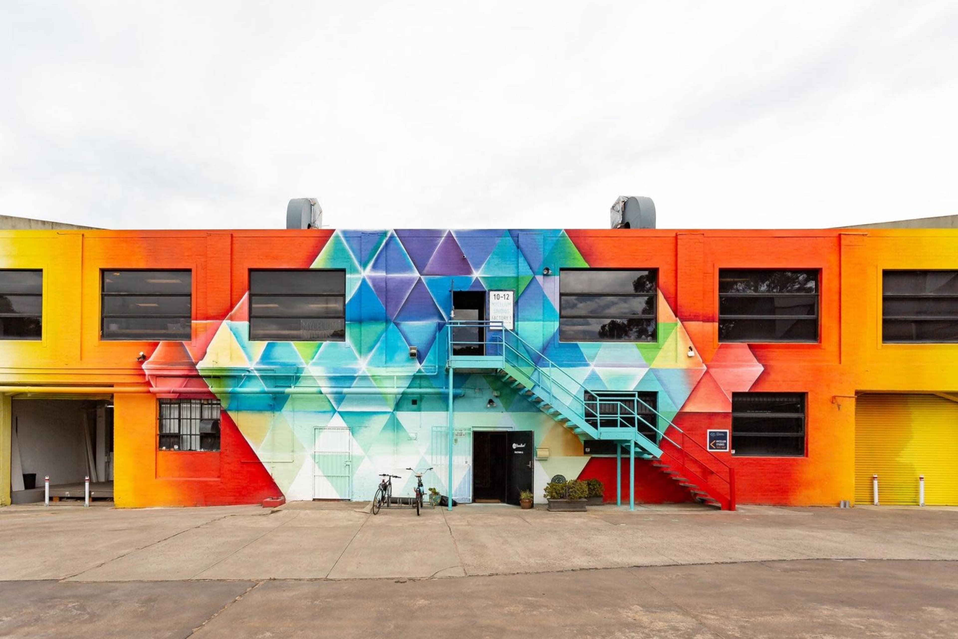 A large, colorful mural featuring geometric patterns adorns the facade of a building with a staircase and bike parked outside.