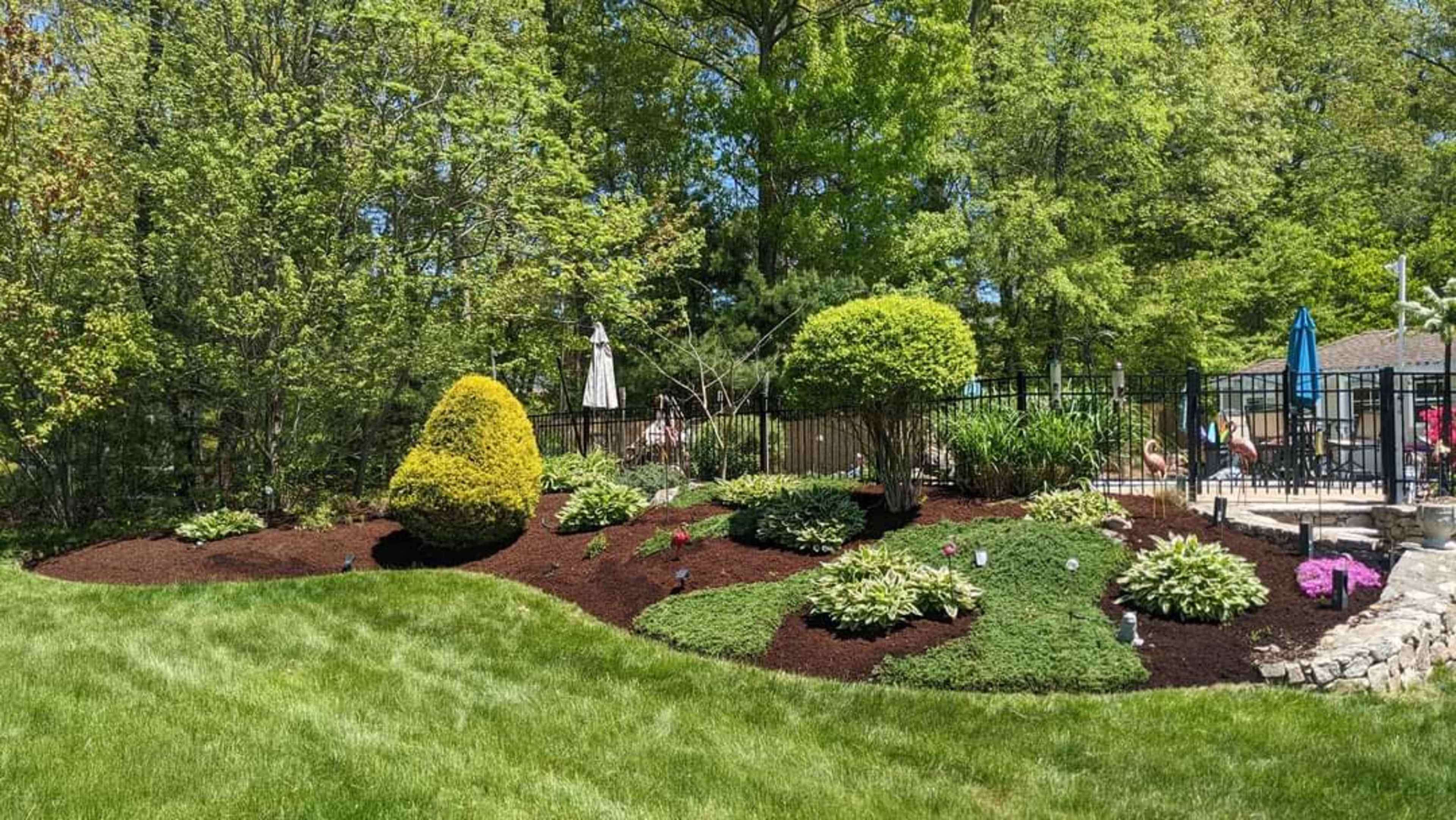 The image shows a landscaped garden featuring manicured bushes, colorful flowers, and a swimming pool area enclosed by a black fence.