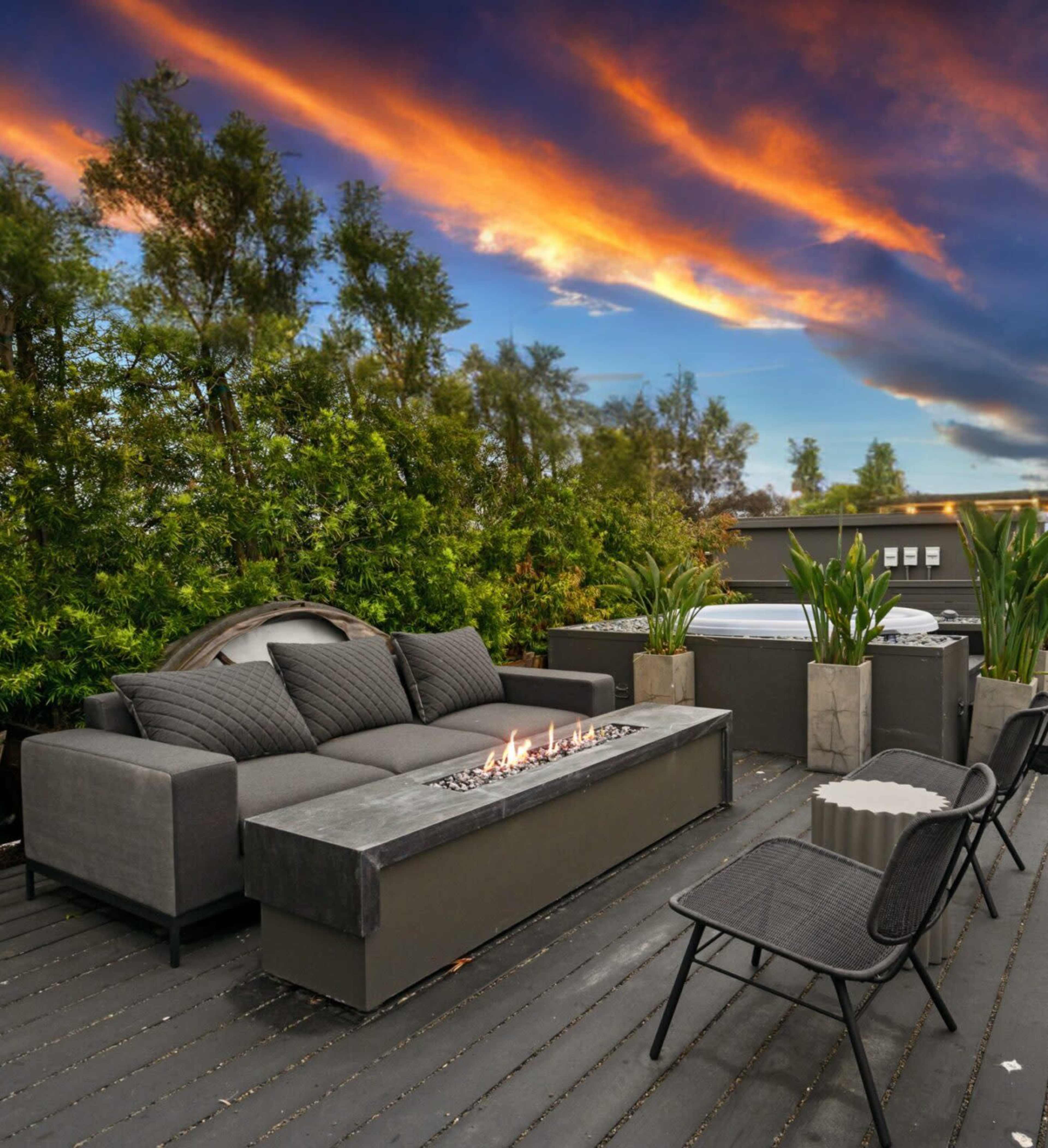 A modern outdoor seating area features a fire table, comfortable sofa, and planters against a backdrop of a vibrant sunset sky.