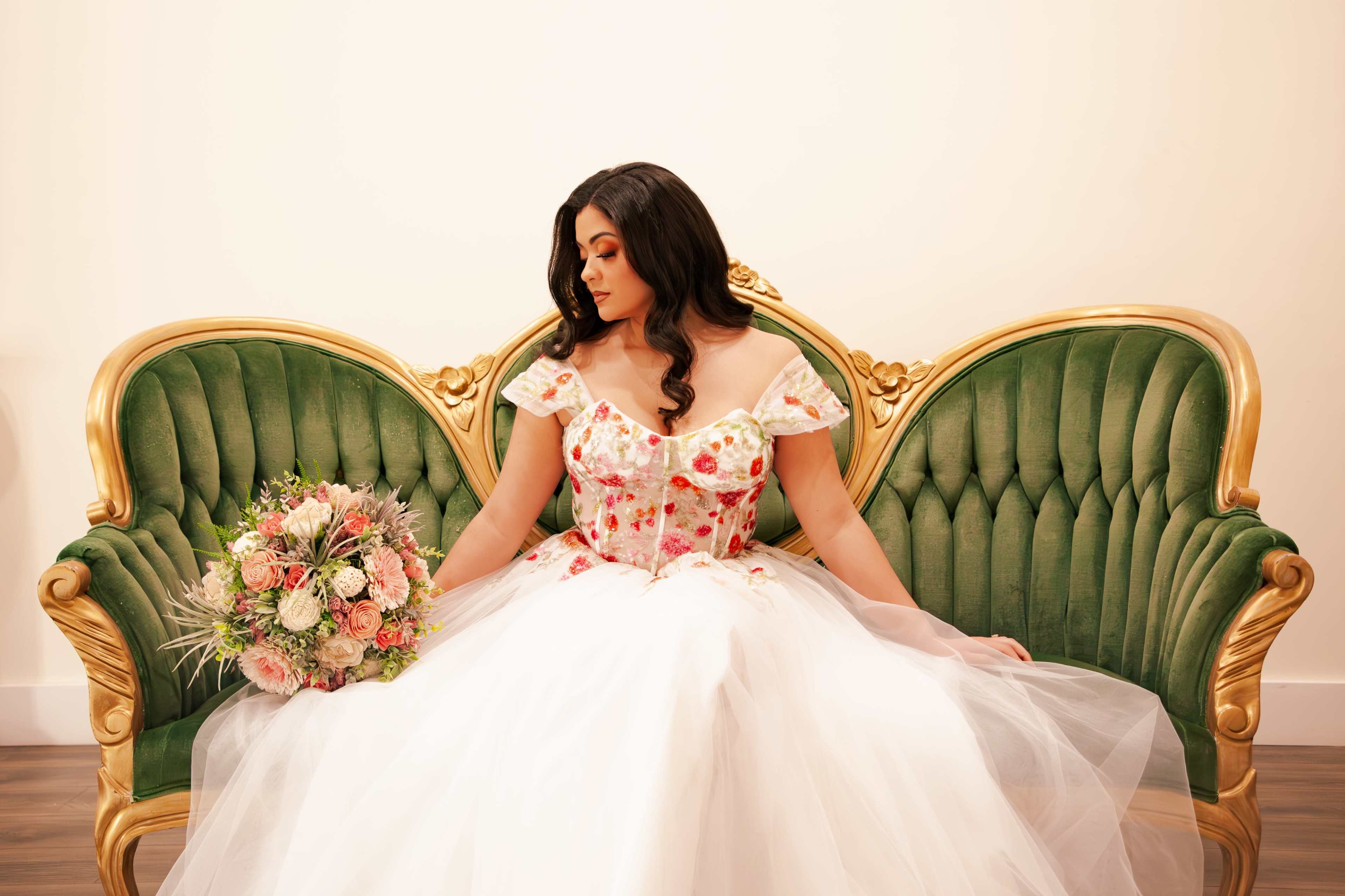 A woman in a floral gown sits on an elegant green velvet sofa, holding a bouquet of flowers.