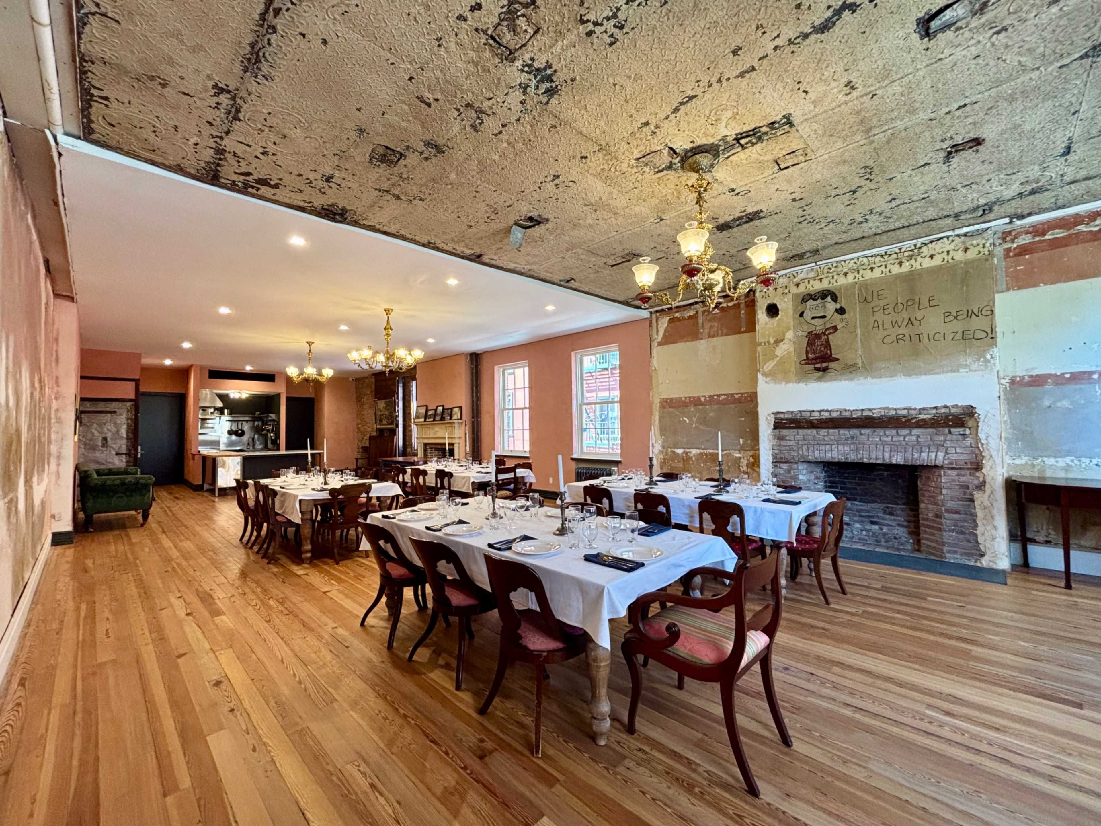The image shows a dining room set for a meal, featuring tables with white linens, wooden chairs, and a fireplace, along with exposed brick and vintage chandeliers.