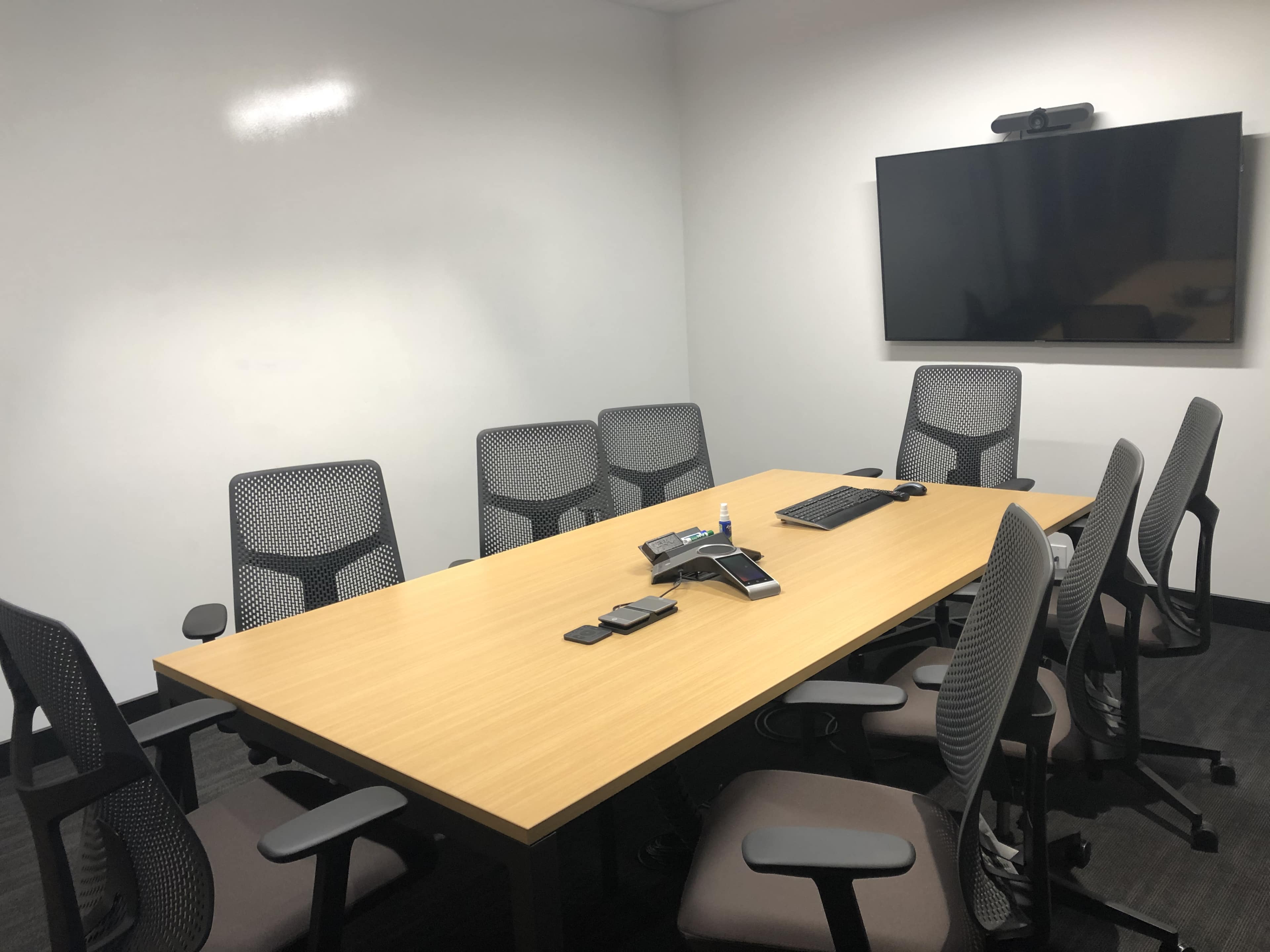 A conference room is set up with a large wooden table surrounded by eight mesh office chairs, equipped with a phone and a monitor on the wall.