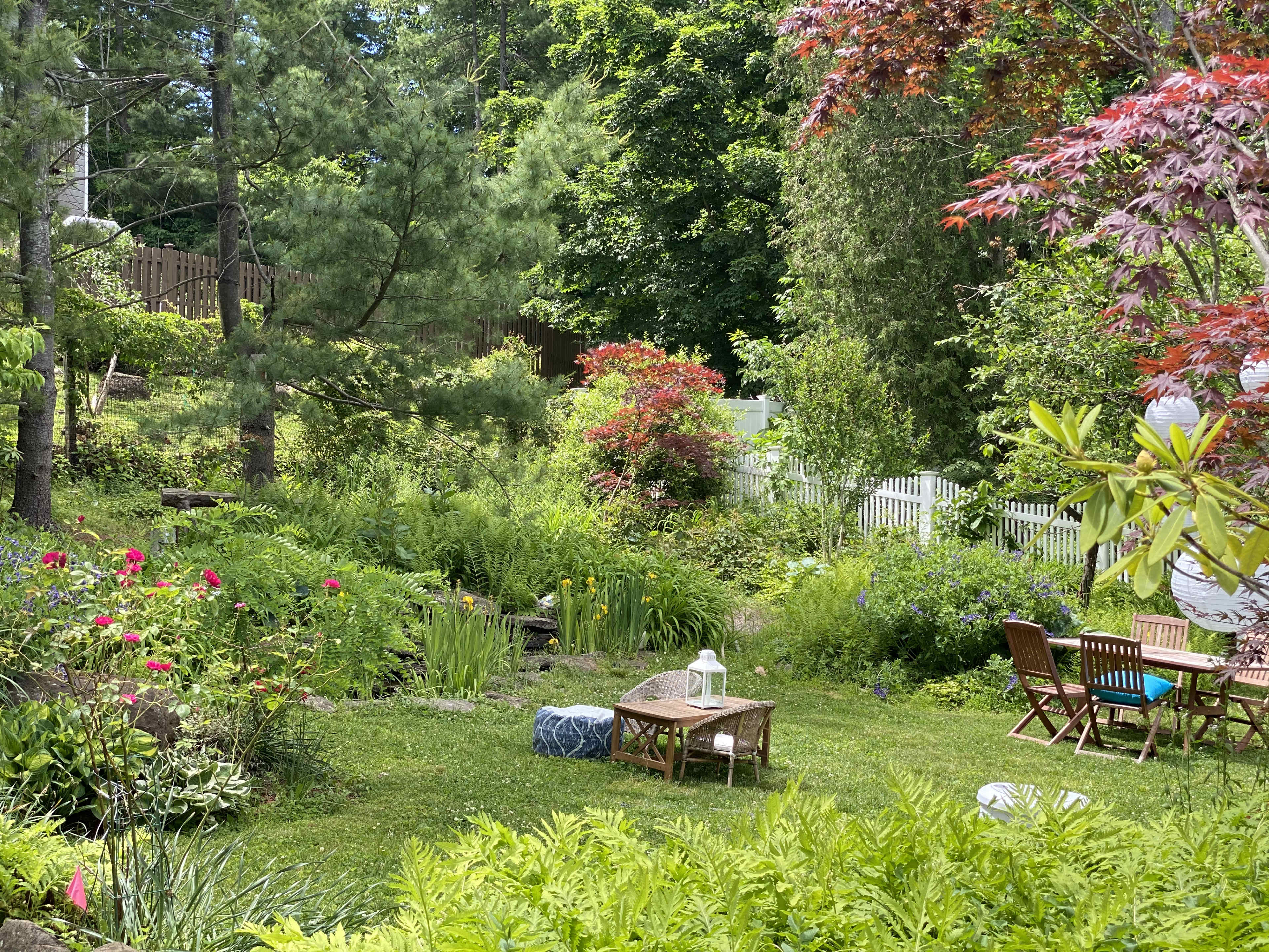 A lush garden features various plants, flowers, and a seating area with tables and chairs amidst greenery.