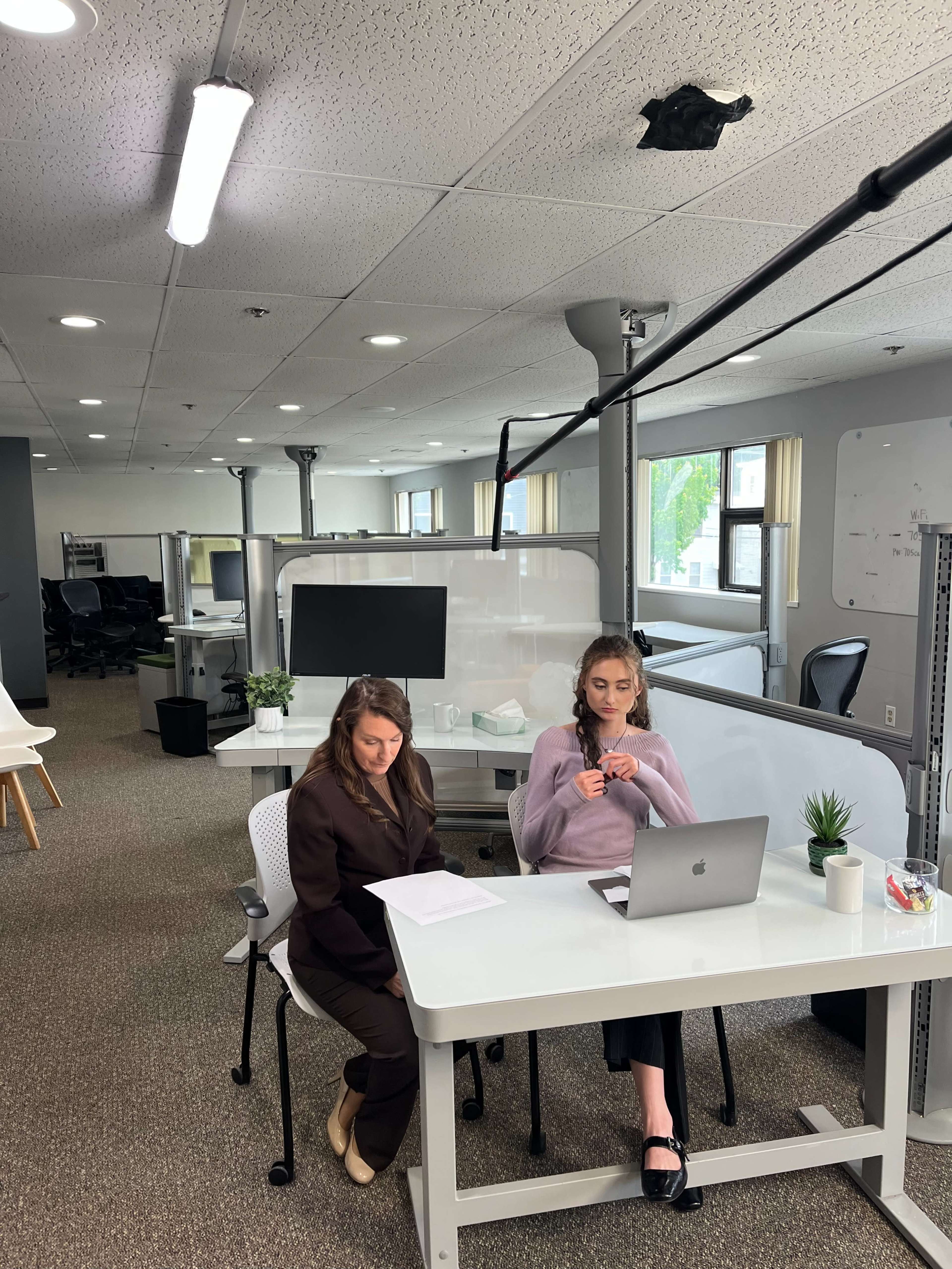 Two women are seated at a modern office workspace, one reviewing documents while the other works on a laptop.