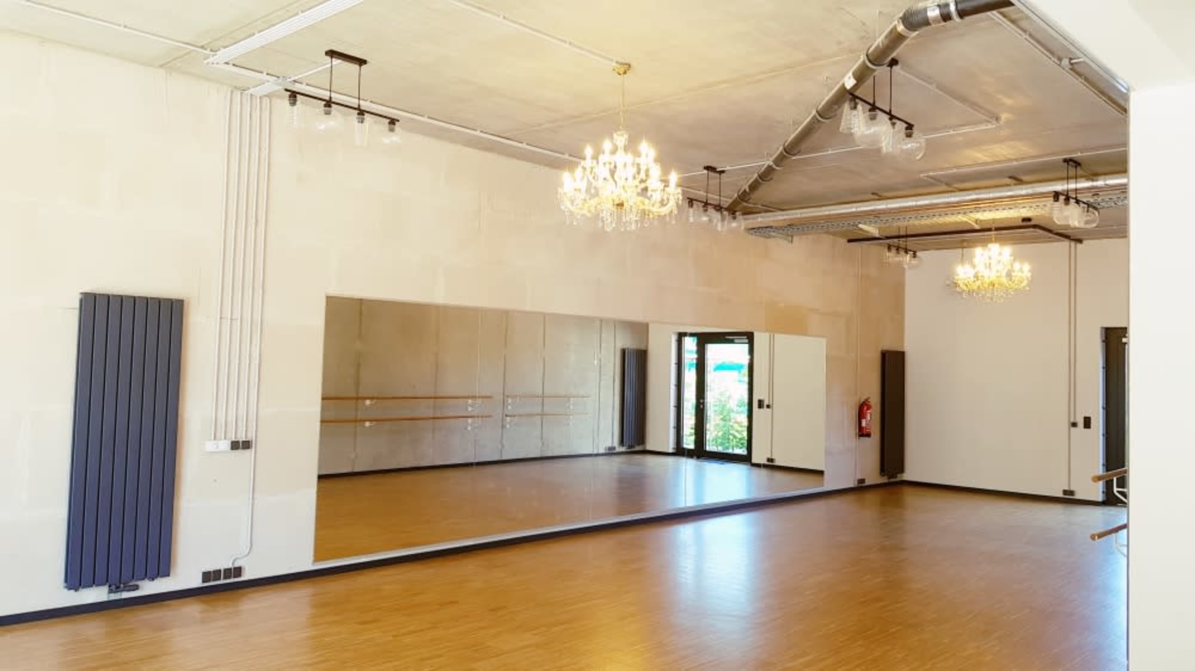 A bright, spacious dance studio featuring wooden flooring, large mirrors, and elegant chandeliers.