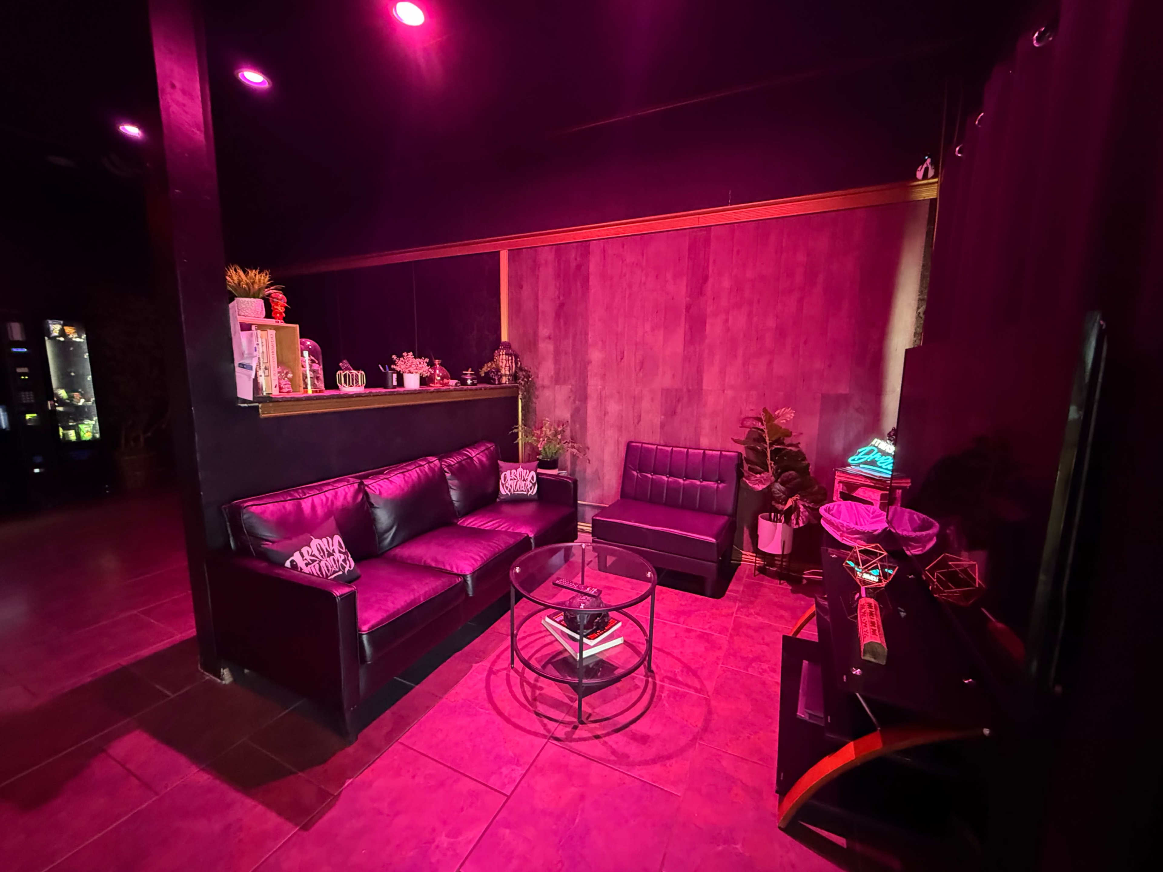 The image shows a dimly lit lounge area with black sofas, a glass coffee table, and decorative plants, illuminated by pink lighting.