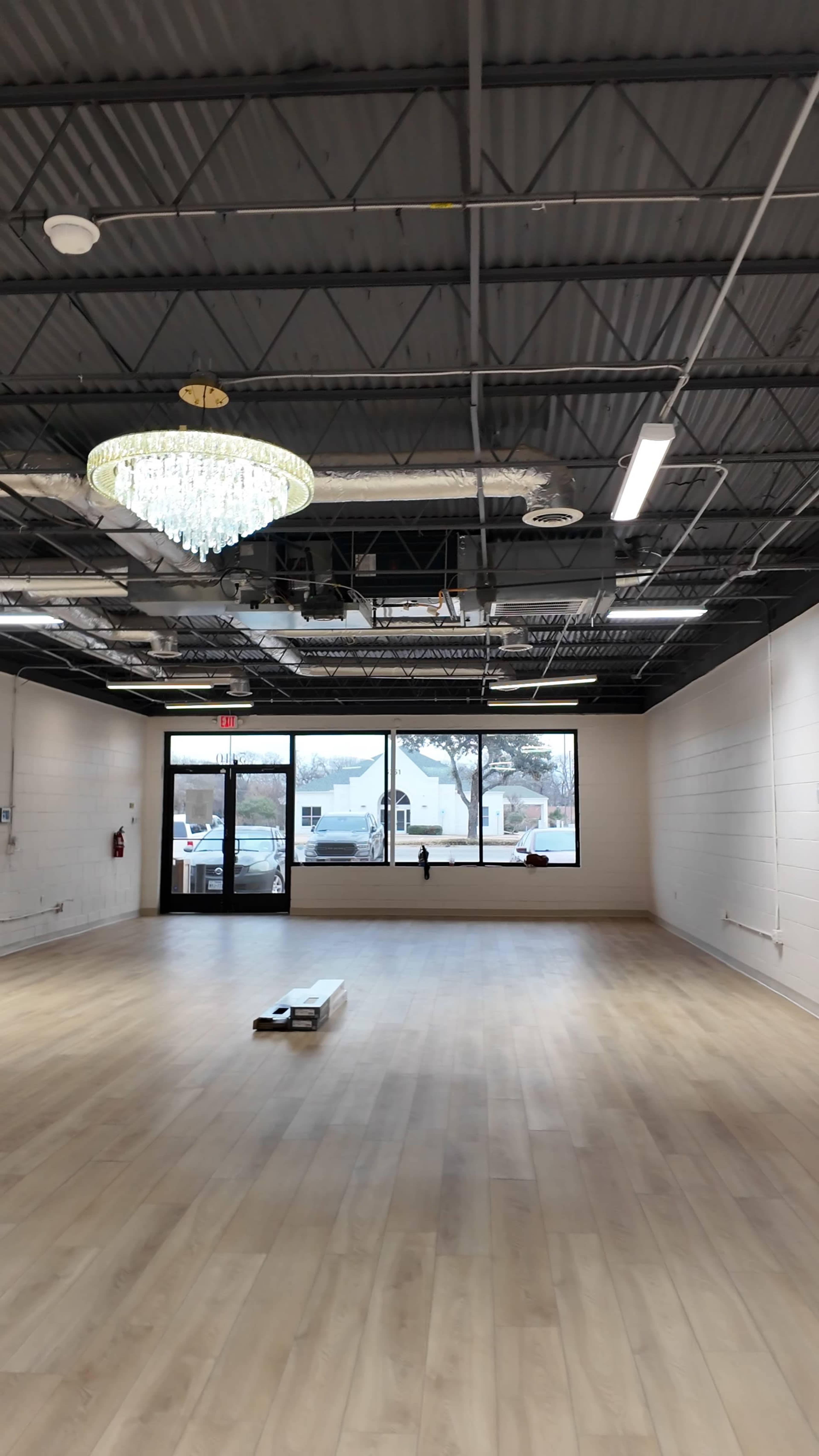 The image shows a spacious, empty room with wooden flooring, a chandelier hanging from the ceiling, and large windows allowing natural light to enter.