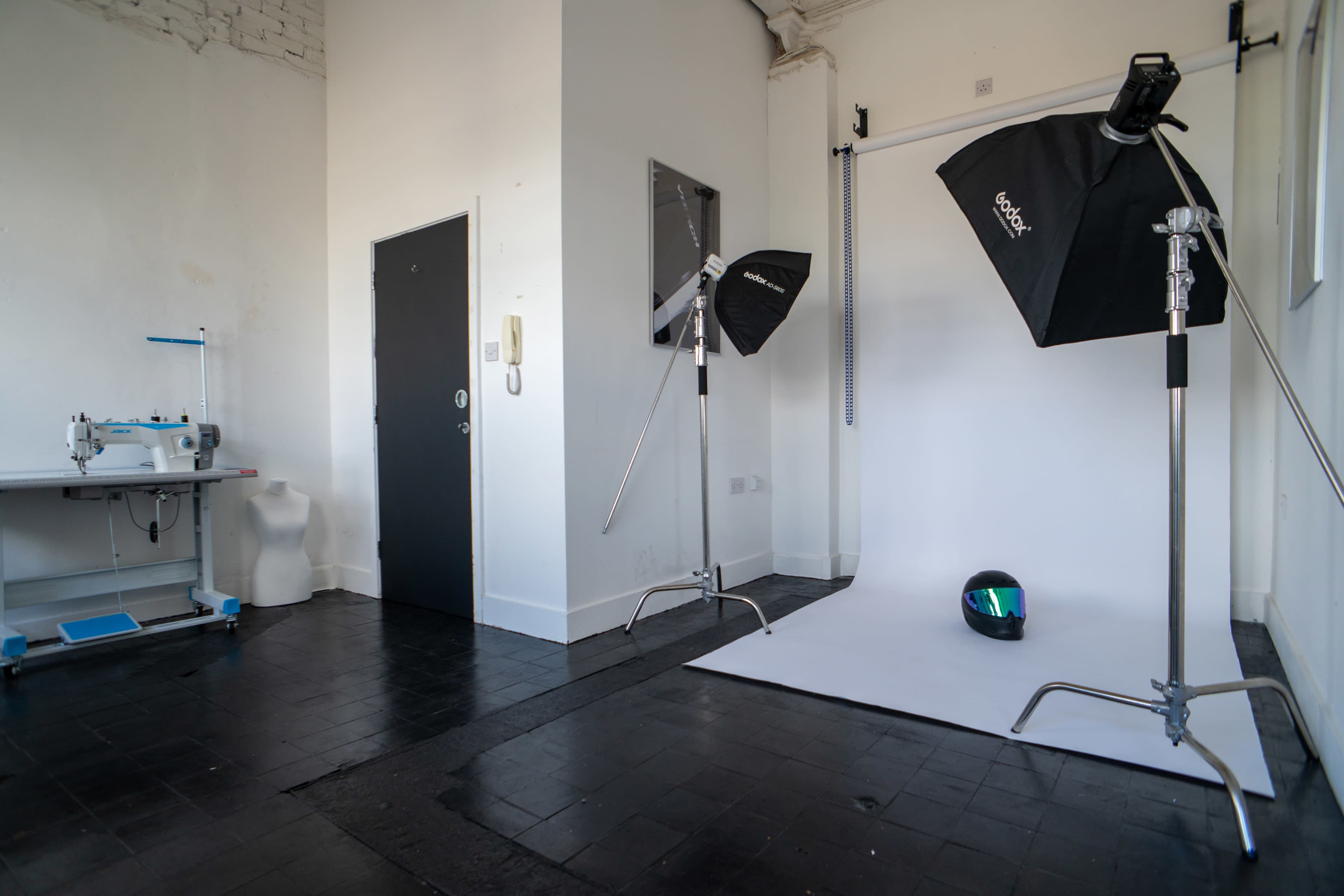 The image shows a photography studio with a sleek white backdrop, two softbox lights, a black helmet, and a sewing machine in the corner.