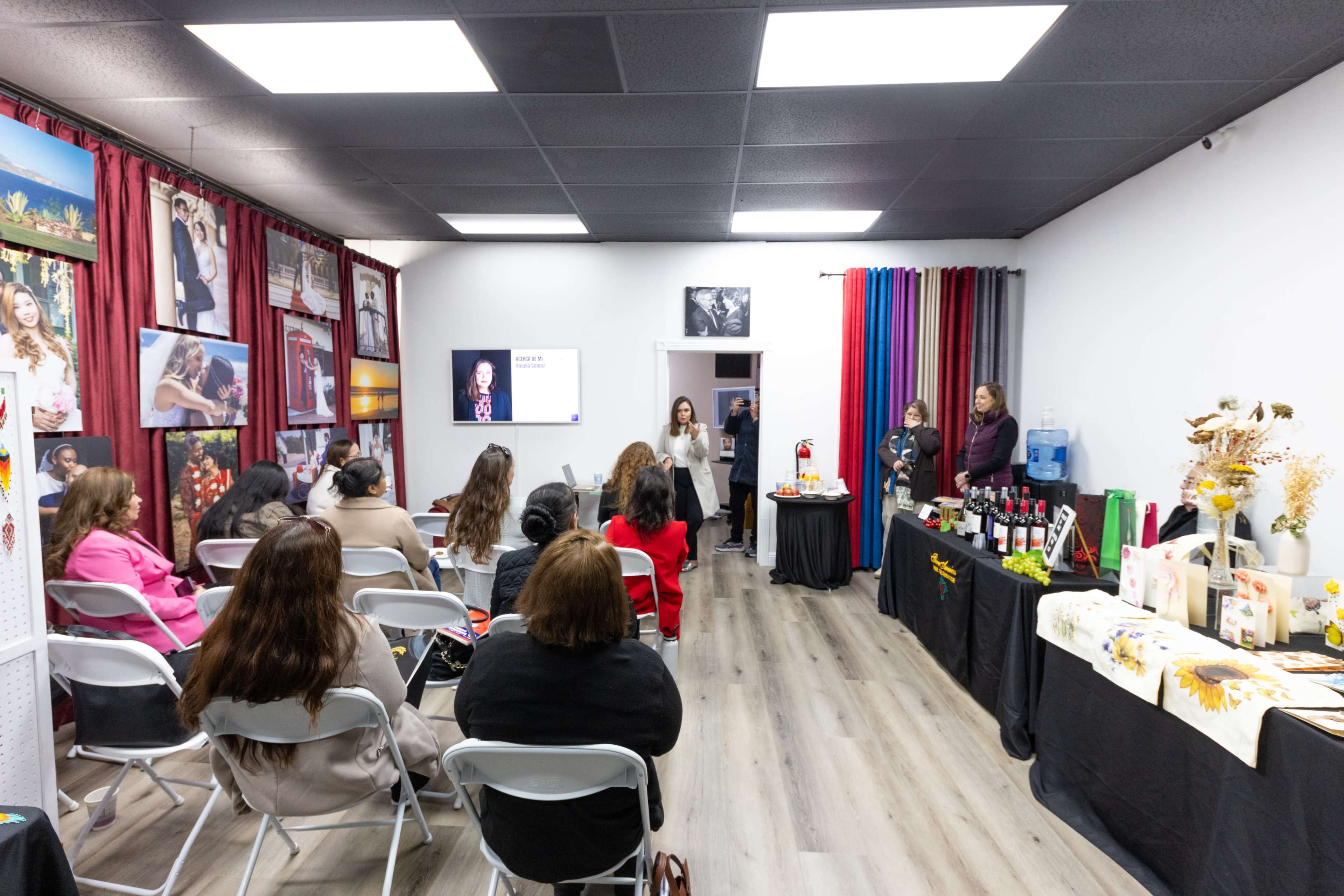 A small group of people sits in chairs facing a presenter in a brightly lit room adorned with photographs and a display table featuring various products.